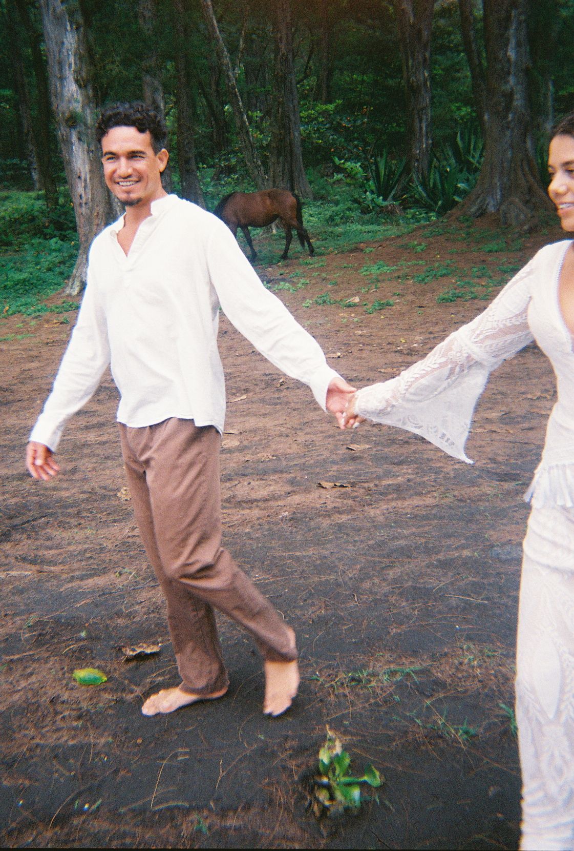 Smiling groom in white shirt and brown pants holding bride's hand while walking barefoot near horses.