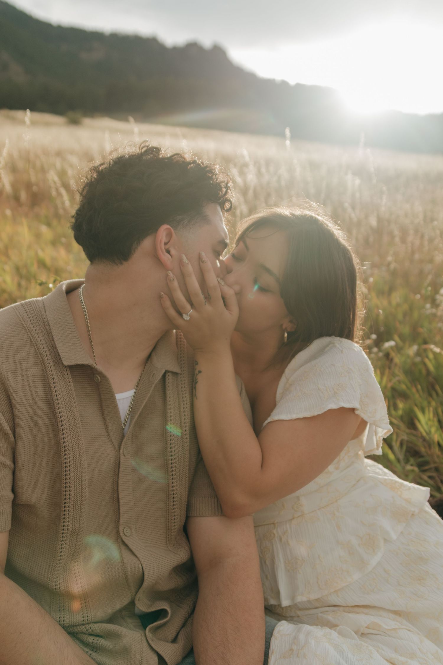 Couple sharing a tender kiss while sitting in a sunlit grassy field with warm golden light.