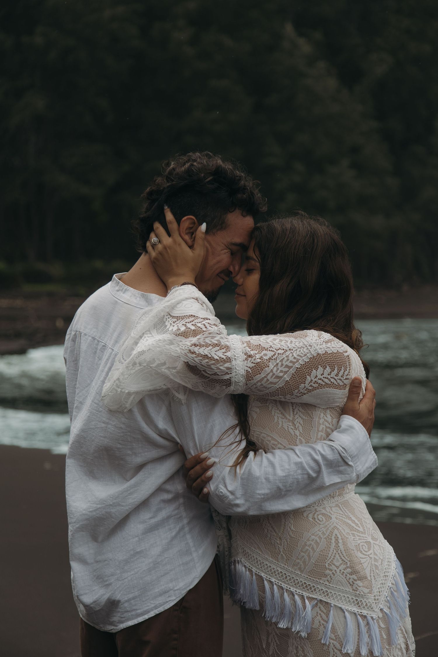 Couple sharing intimate embrace on dark beach with lush forest backdrop during elopement shoot.