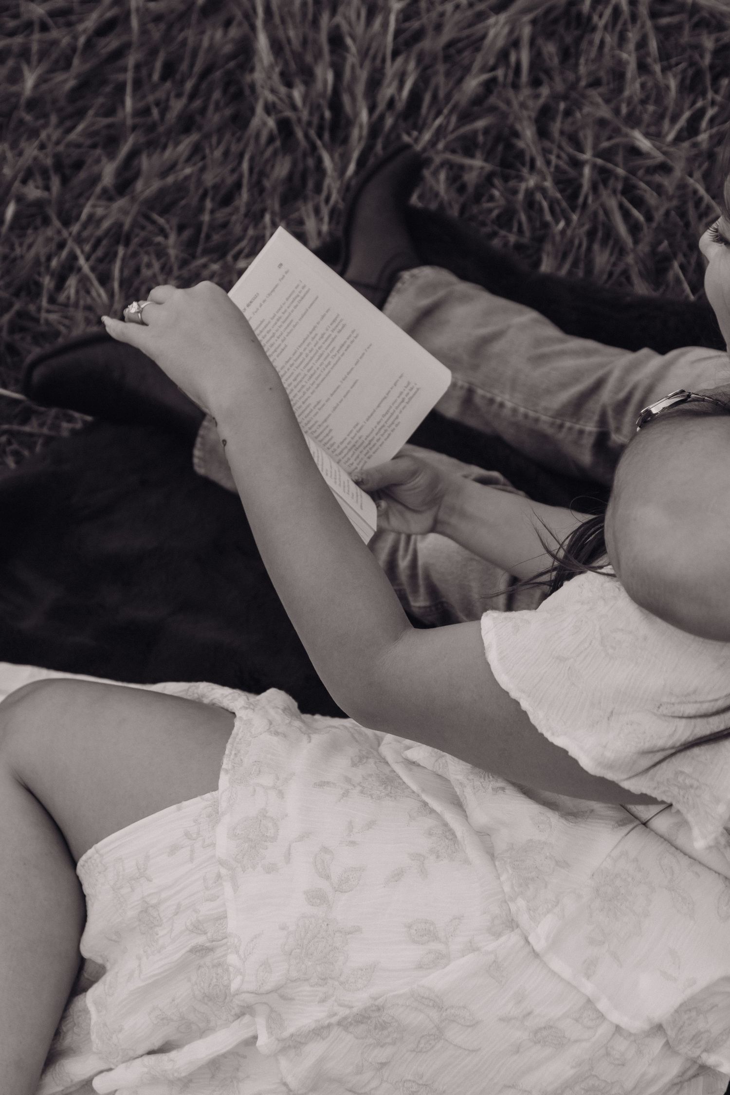 Black and white photo of a person in a floral dress reading a book while lying on a blanket outdoors.