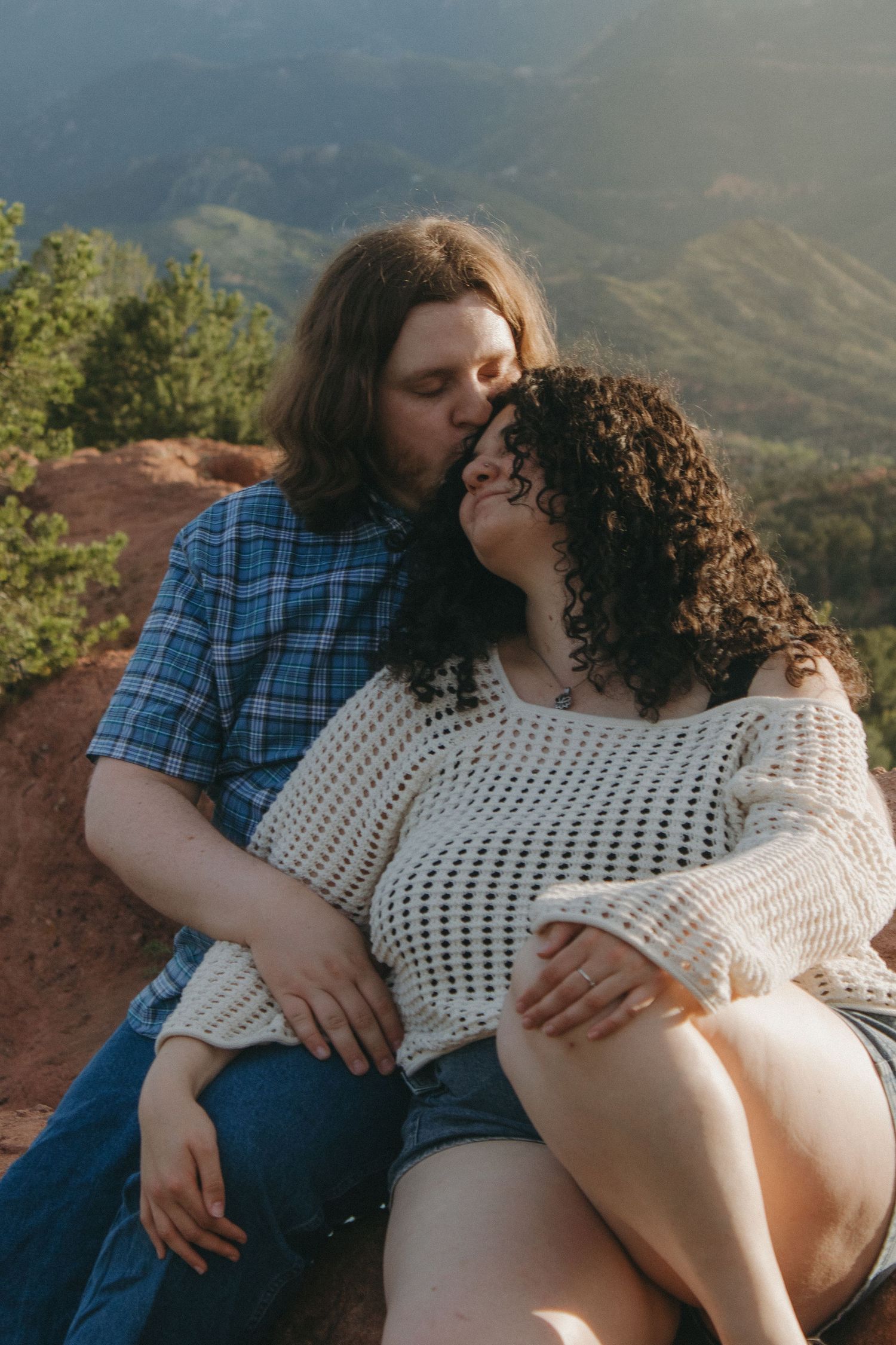 A couple shares an intimate moment while sitting on red rocks with mountain vistas behind them.