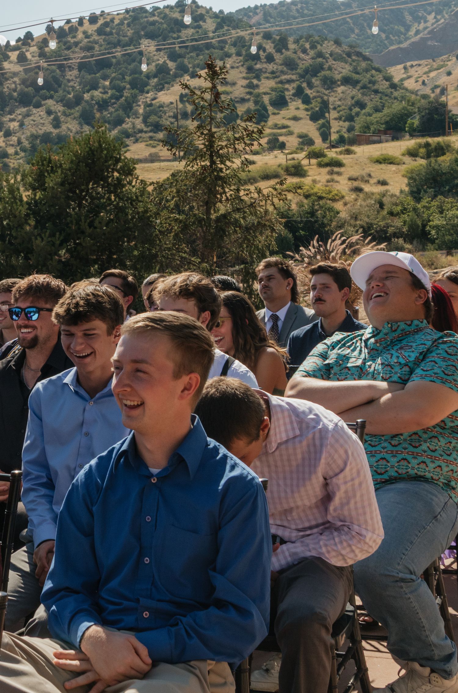 Wedding guests laugh together while seated outdoors with mountains in the background.