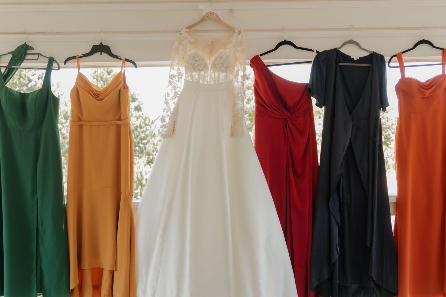 A row of colorful bridesmaid dresses in green, gold, white, red, black and orange hanging on wooden hangers.