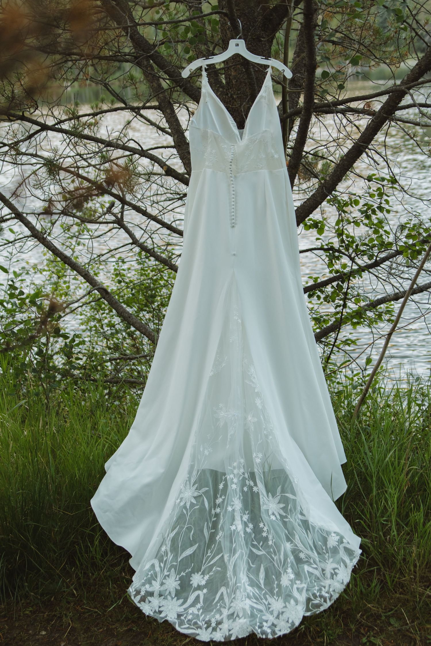 White lace wedding dress hanging on display against natural foliage and water backdrop.
