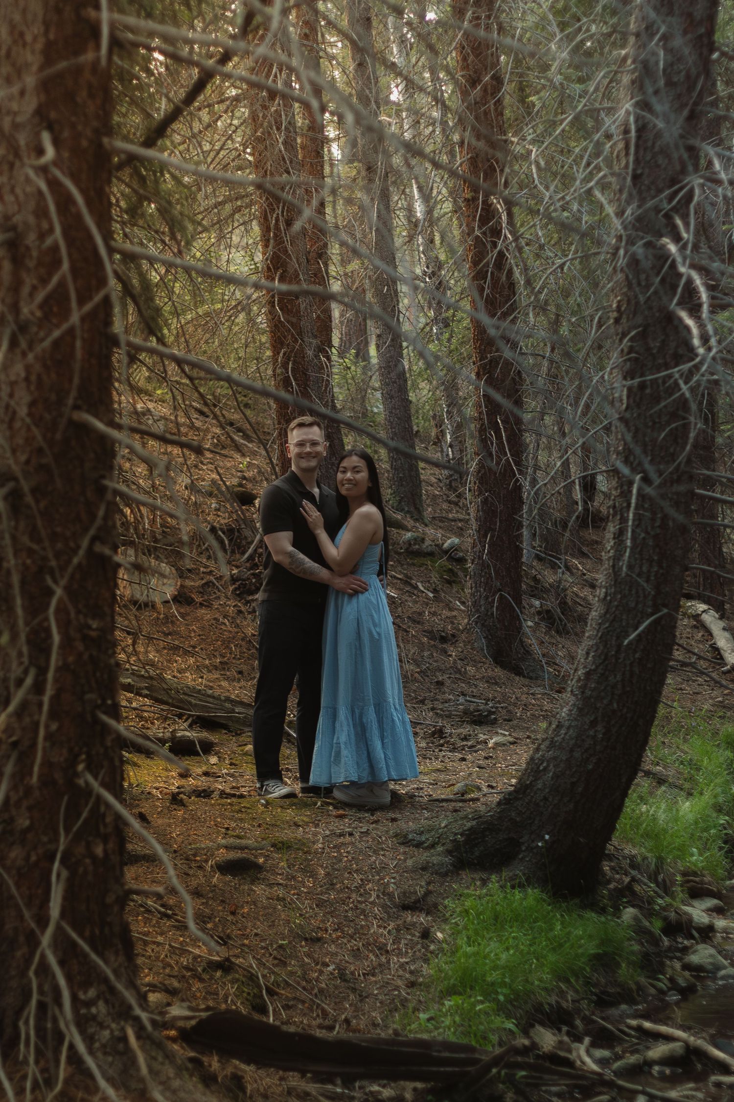 A couple in love poses together on a forest trail wearing black and blue outfits.