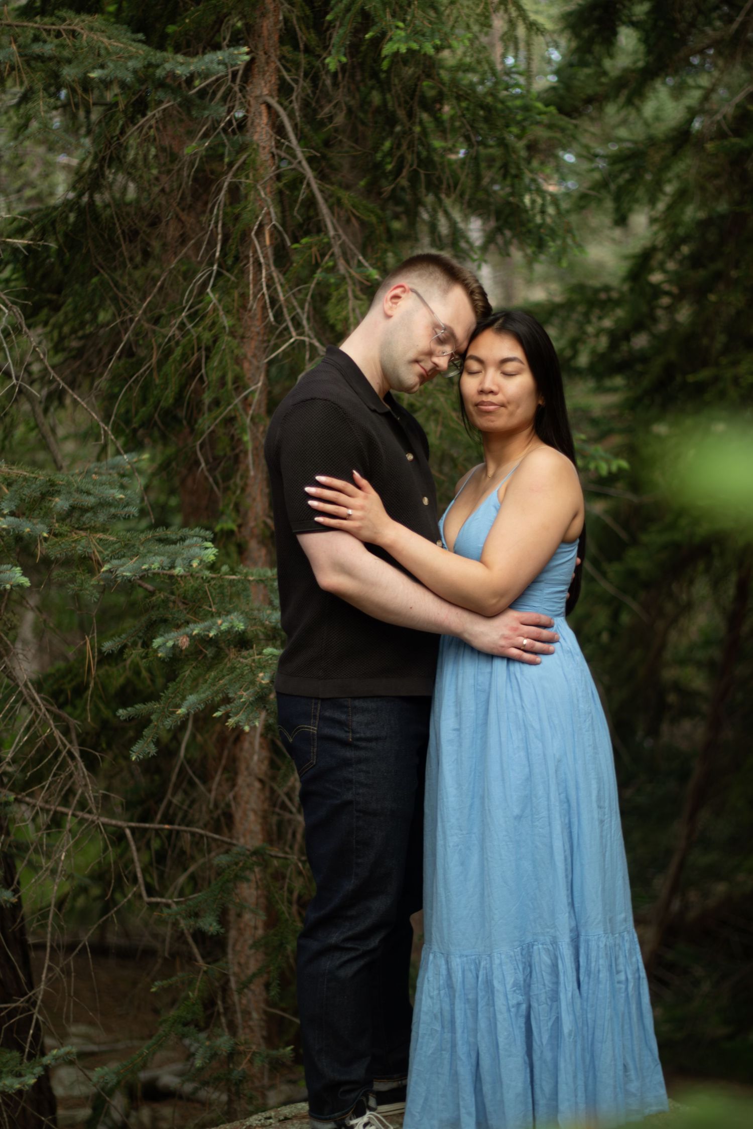 A romantic couple embraces in a forest setting, with her wearing a flowing light blue dress against evergreen trees.