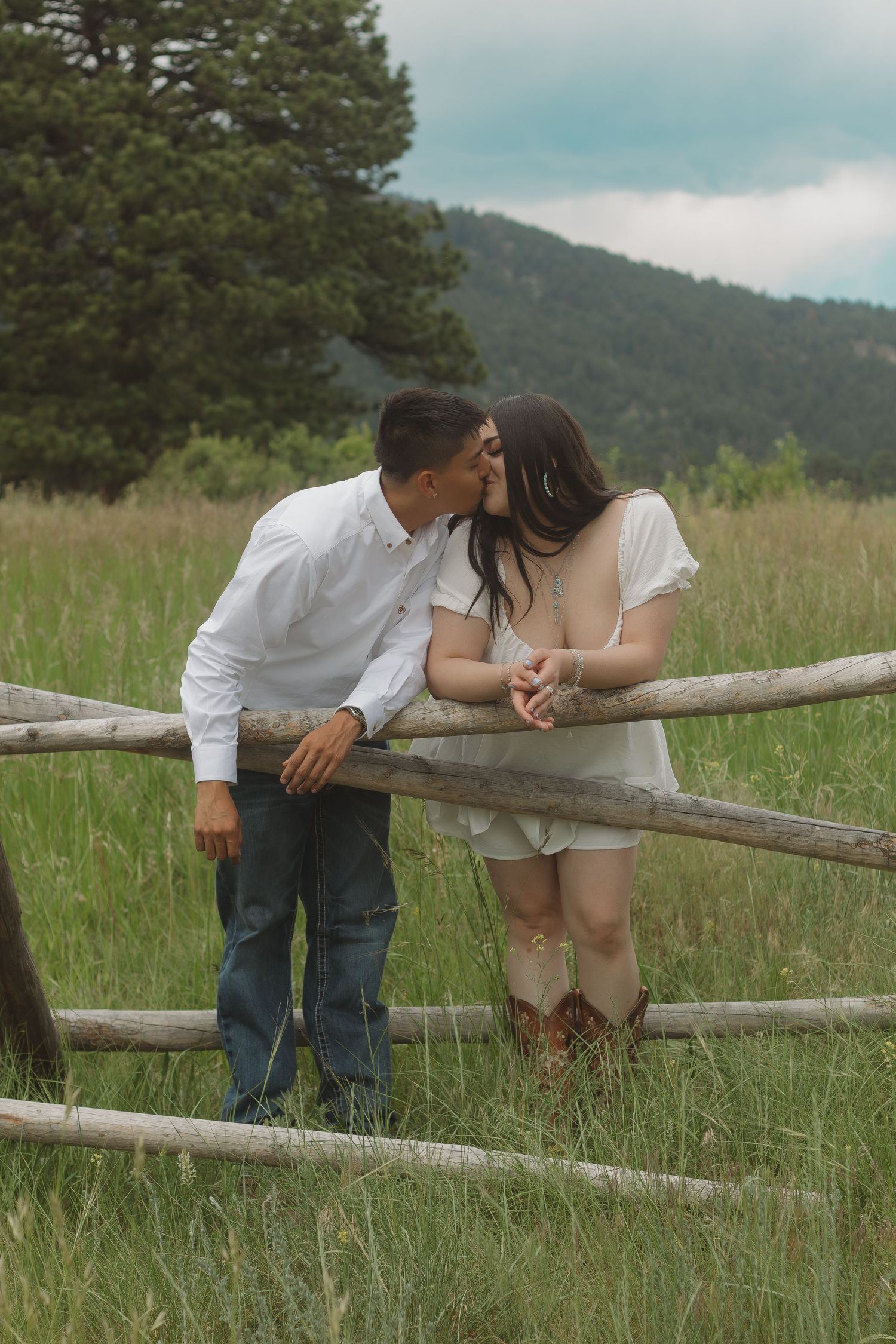 Romantic couple sharing an intimate moment by a rustic wooden fence in a mountain meadow with cowboy boots.