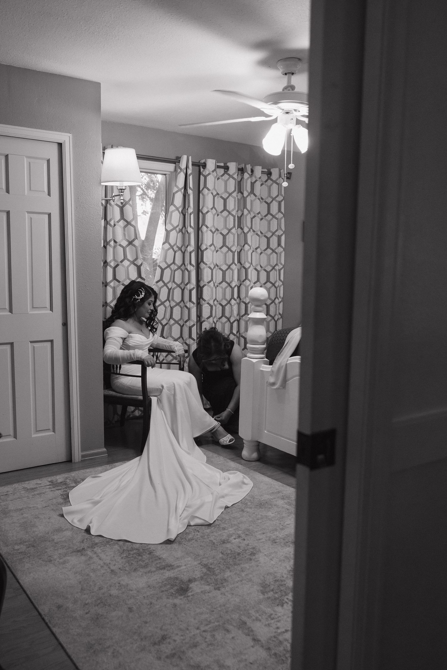 Bride in white wedding gown being helped with shoes in dimly lit room.