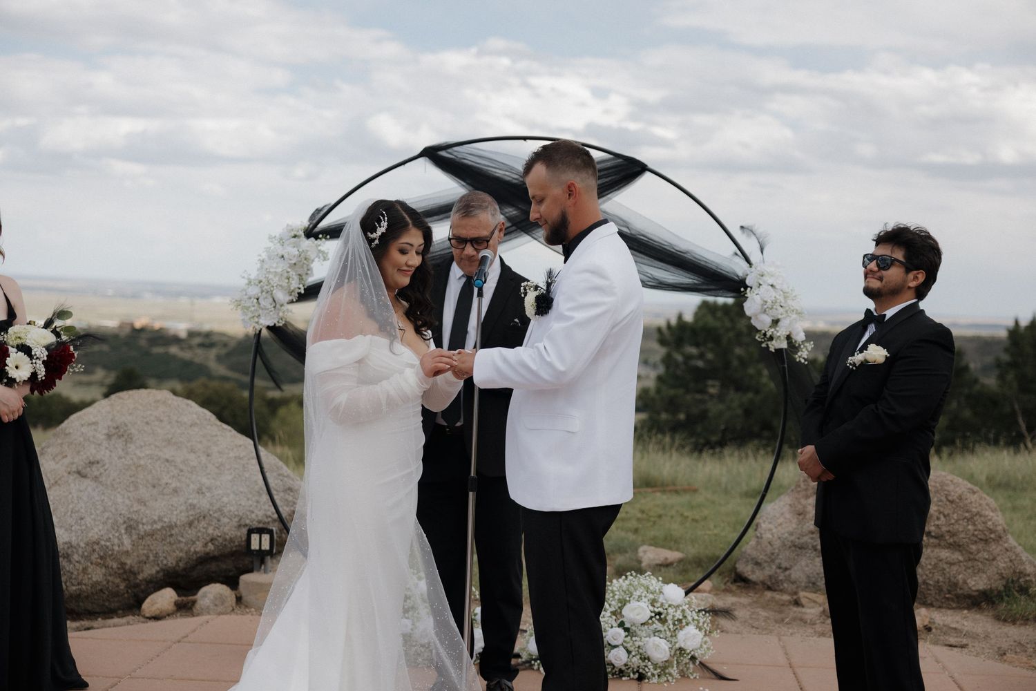 Bride and groom exchange vows outdoors under a circular arch with white flowers at a scenic hilltop wedding ceremony.