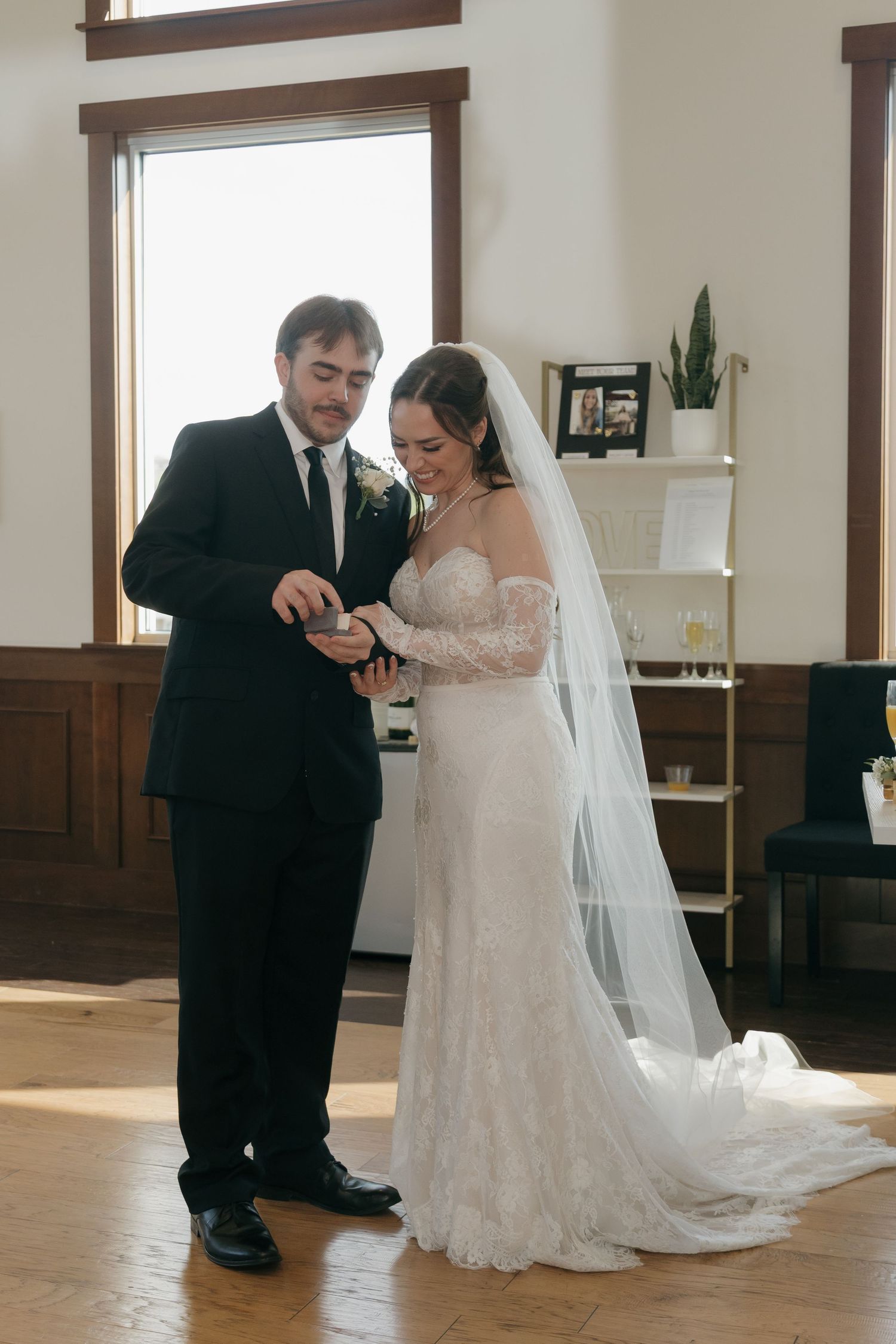 Bride and groom exchanging rings indoors during intimate wedding ceremony, bride wearing lace gown with veil.