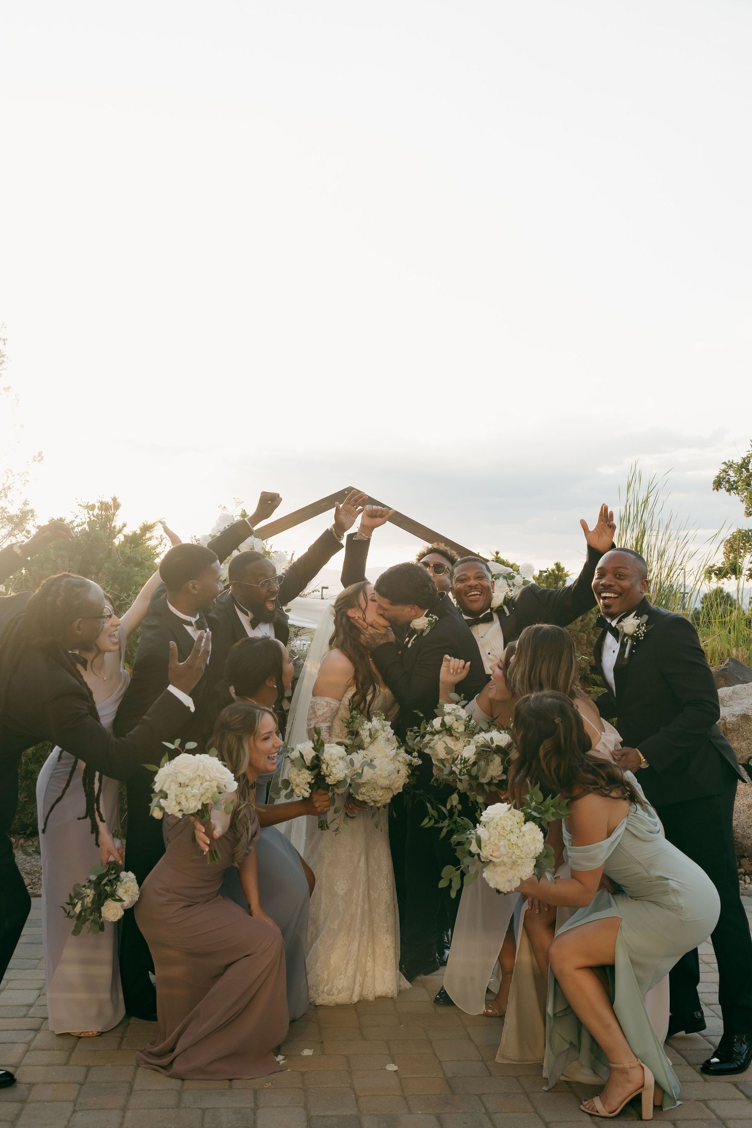 Wedding party surrounding kissing bride and groom in golden sunset light with white bouquets.