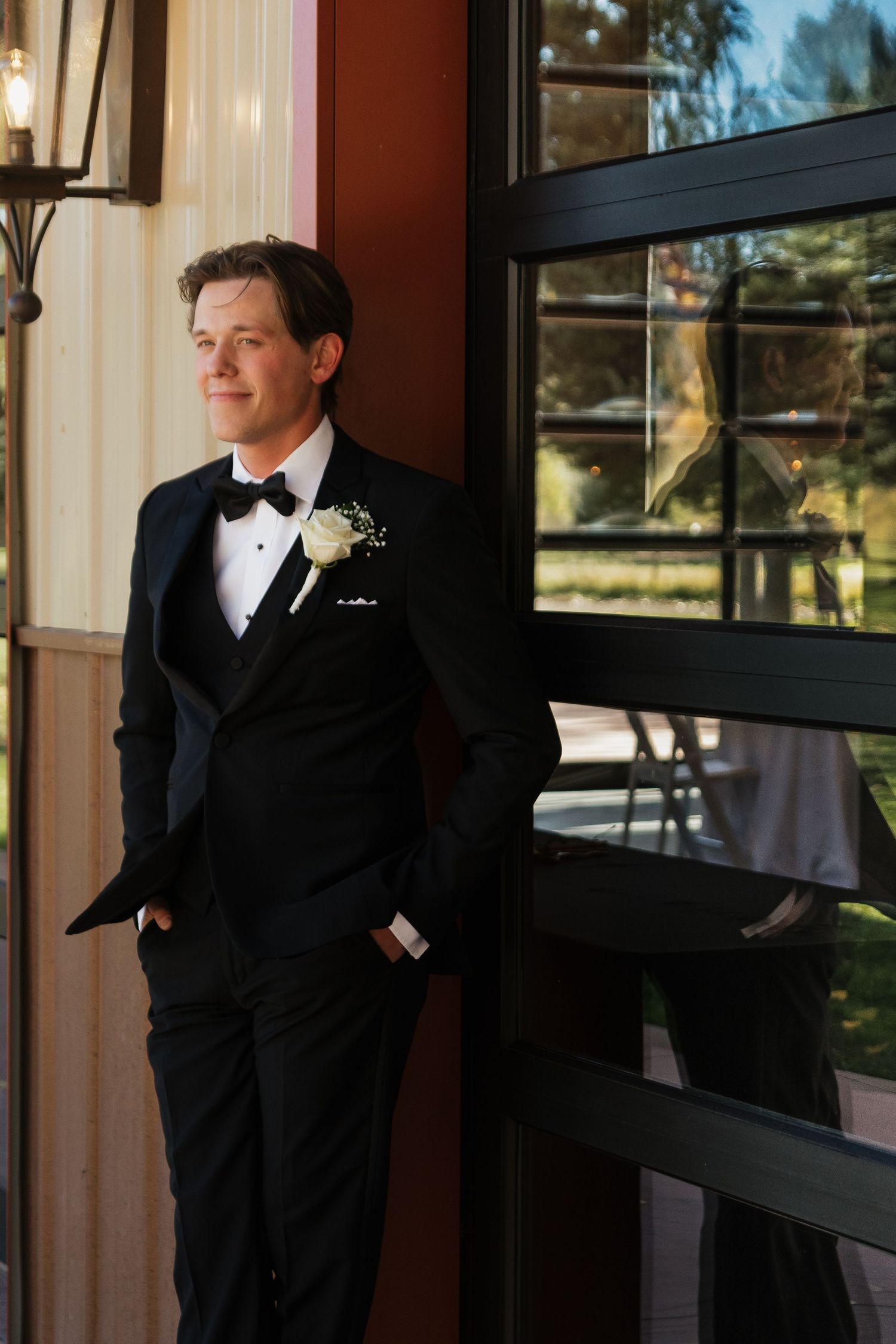 Groomsman in classic black tuxedo poses by window in elegant venue.