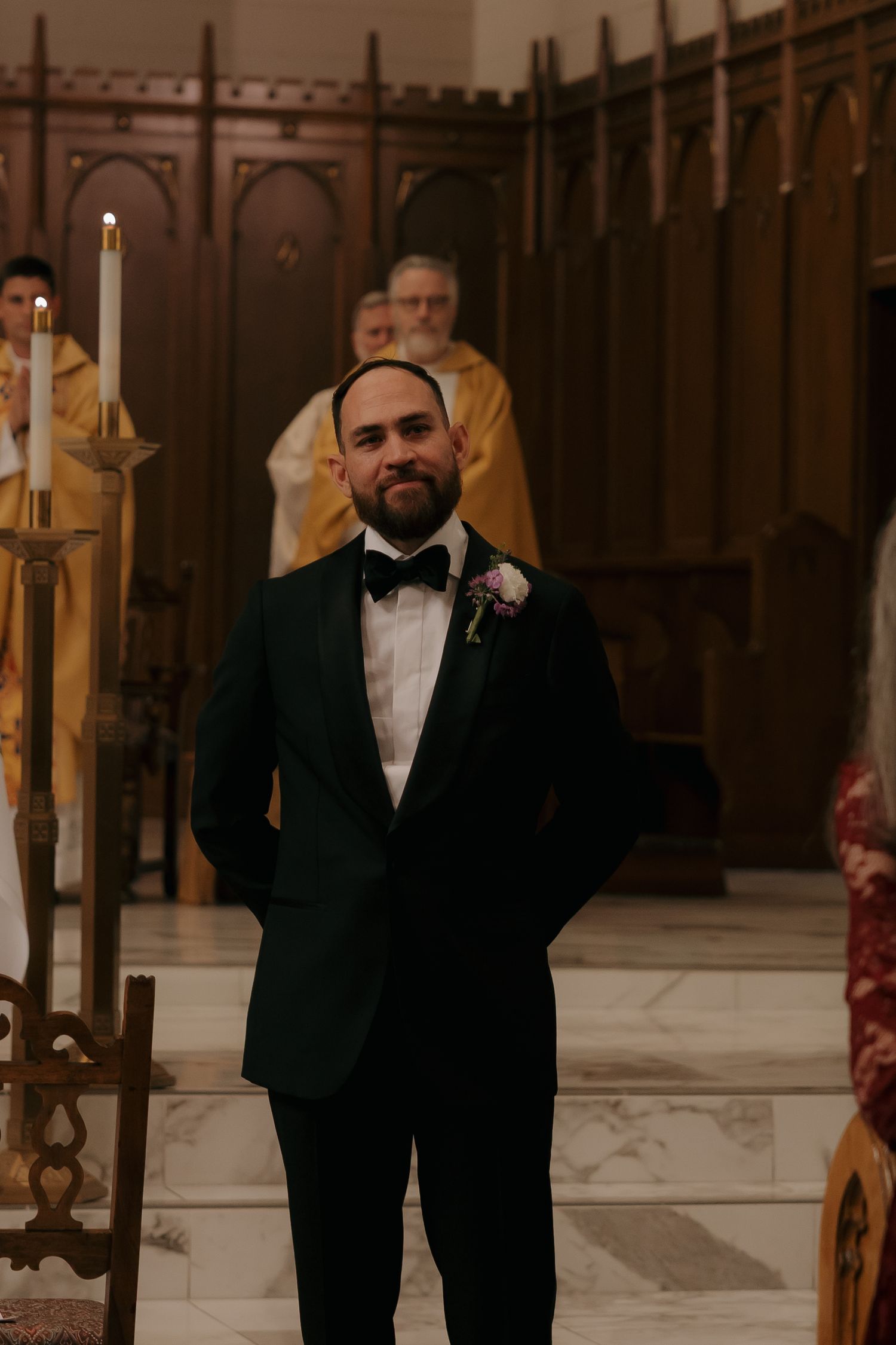 Groom in dark tuxedo with boutonniere stands at church altar during wedding ceremony with officiant in background.