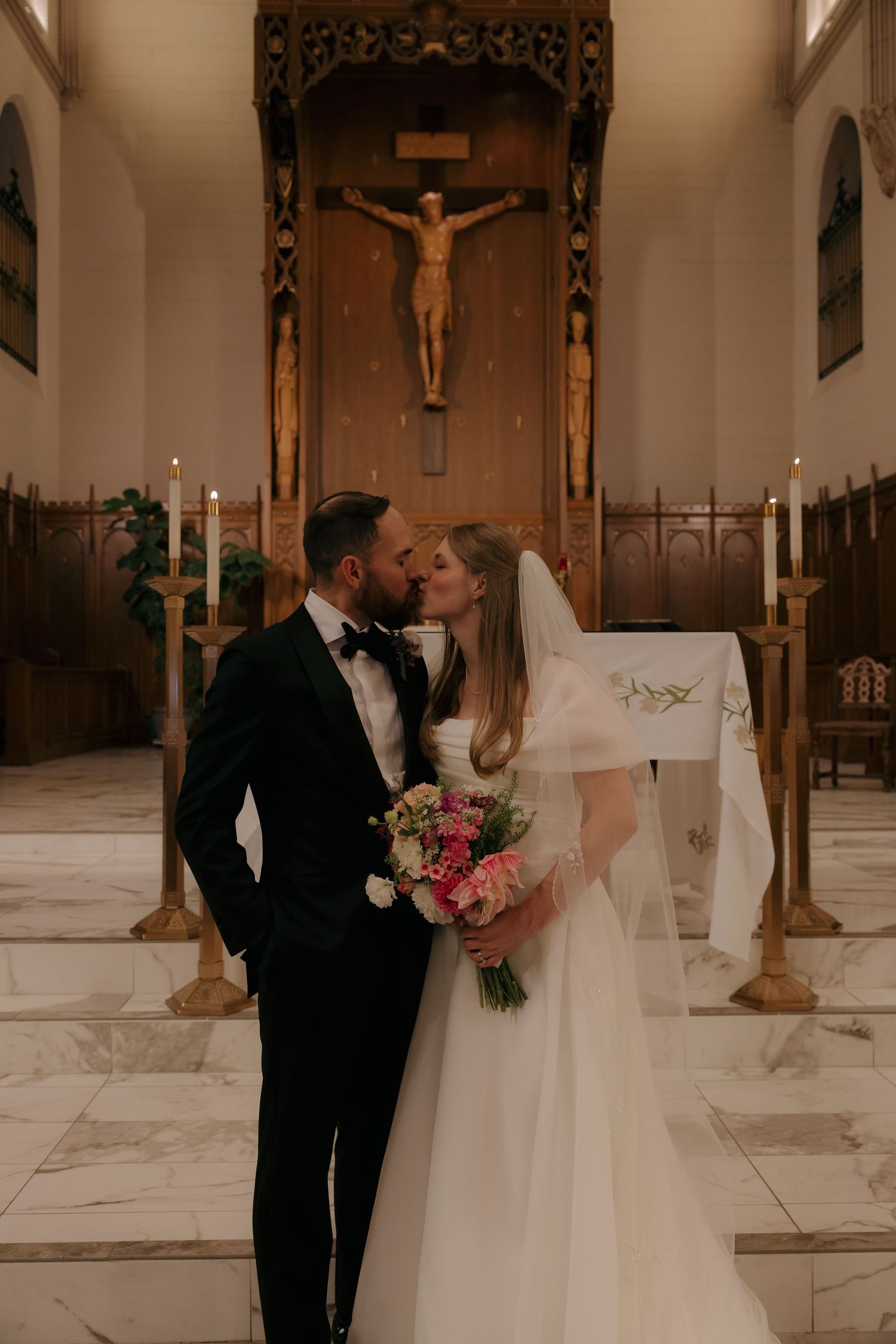Newlyweds share first kiss at church altar beneath crucifix, bride holding pink and white bouquet.