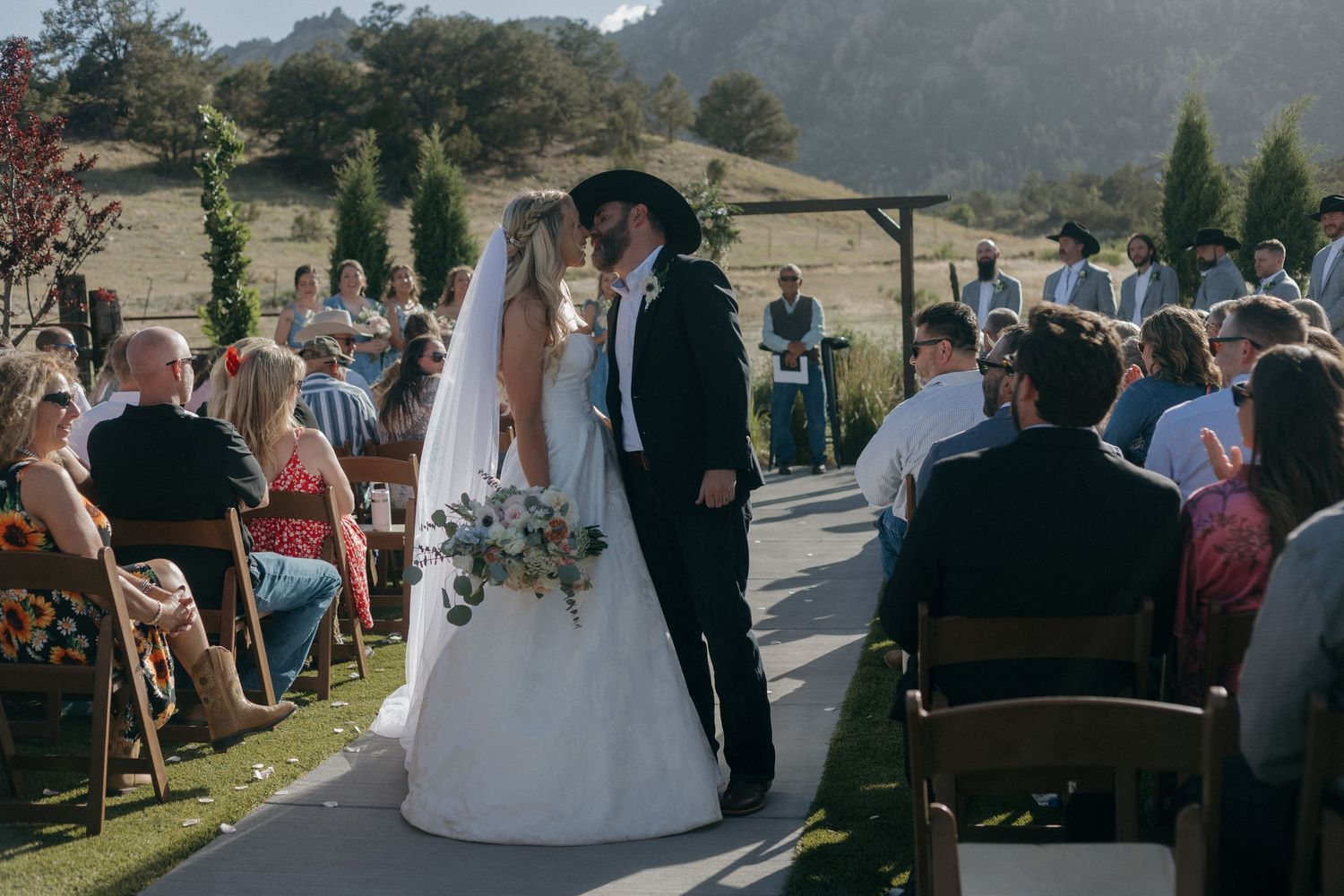 Bride and groom share first kiss at outdoor wedding ceremony with guests seated on both sides amid scenic mountain backdrop.