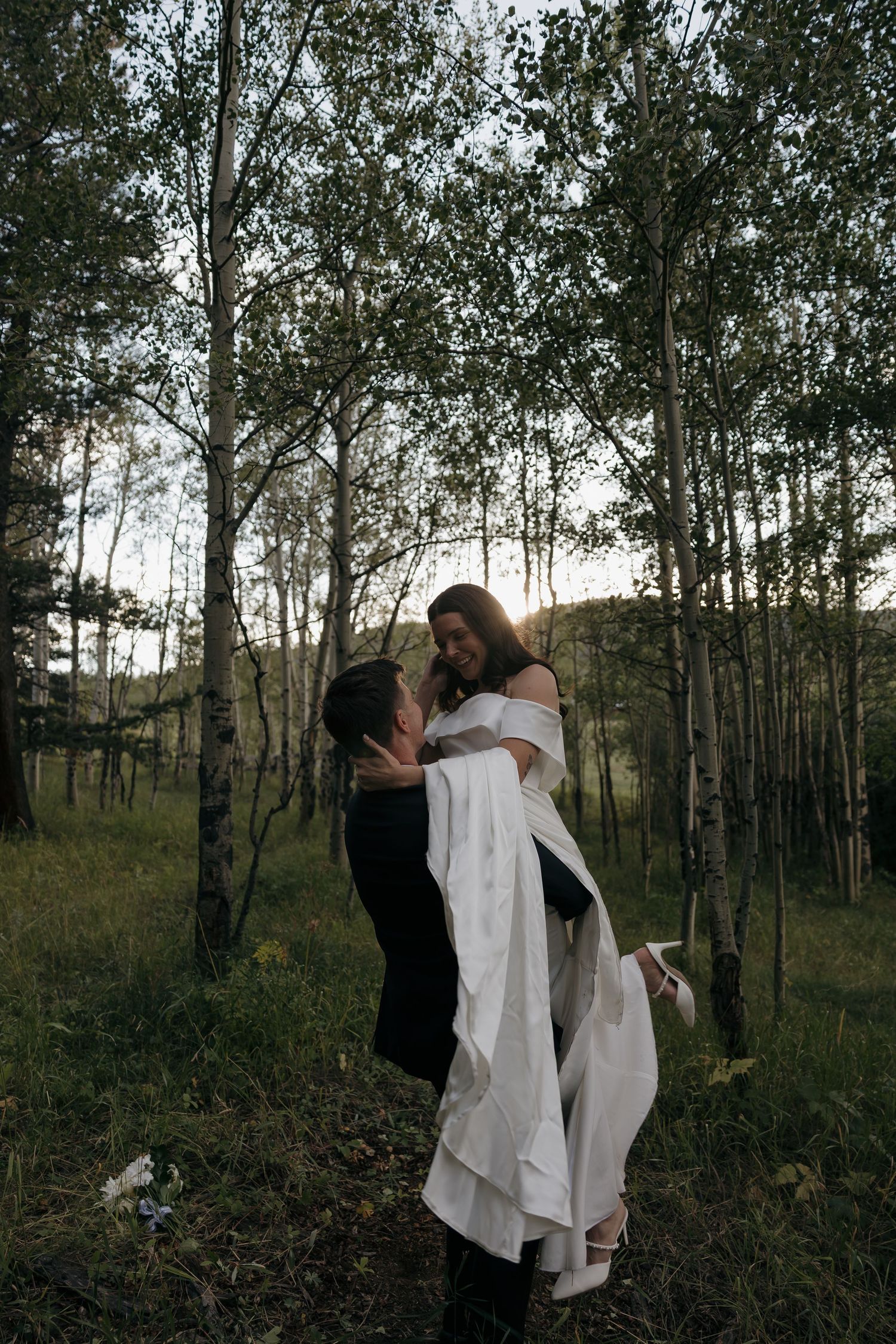 Groom lifting smiling bride in white gown in golden-lit birch tree forest.