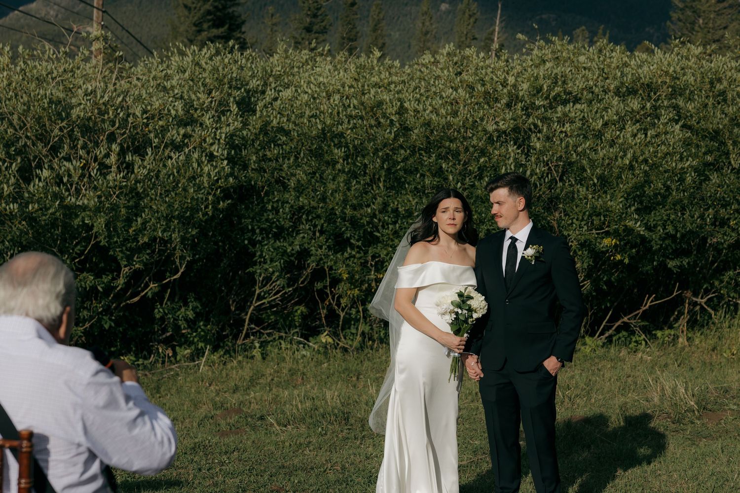 Bride in white off-shoulder gown and groom in dark suit stand outdoors during wedding ceremony with lush green hedge backdrop.