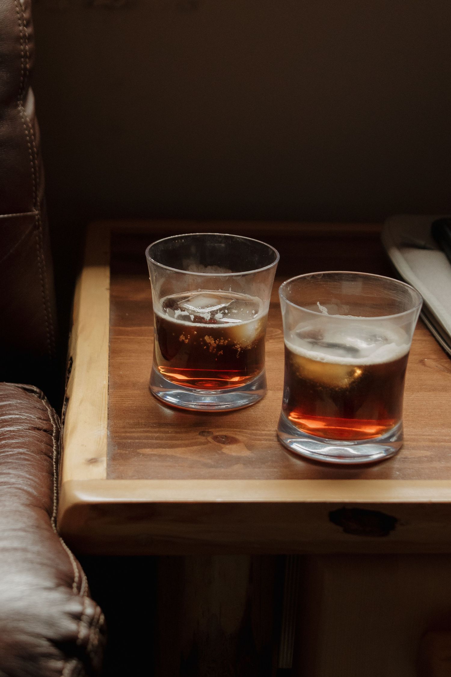 Two glasses of dark liquor with ice on a wooden serving tray next to a leather couch in moody lighting.