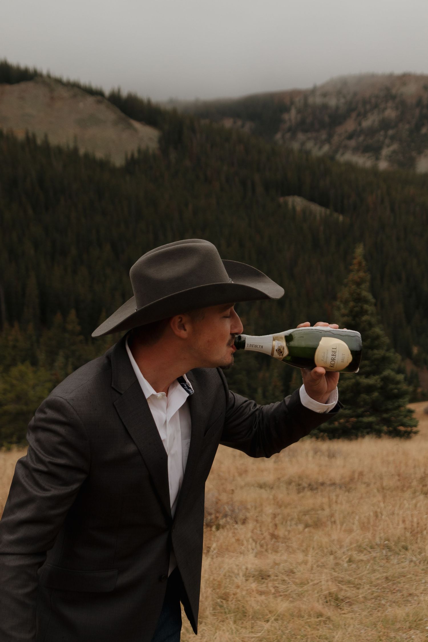 A cowboy in a suit drinks from a bottle against a mountainous forest backdrop.