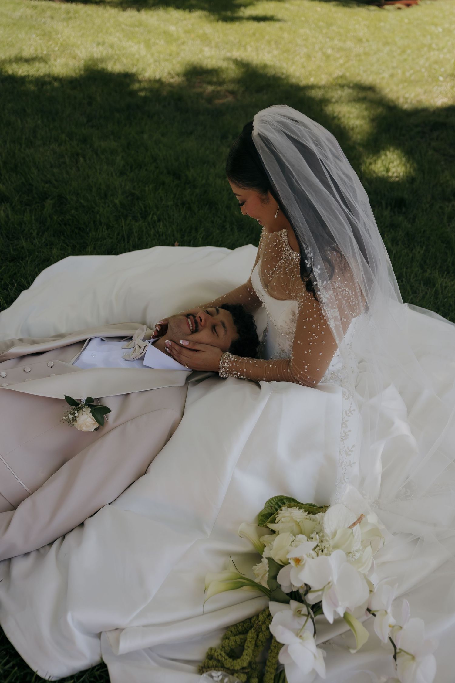Bride in veil tenderly holds grooms face as he lays on her wedding dress outdoors.