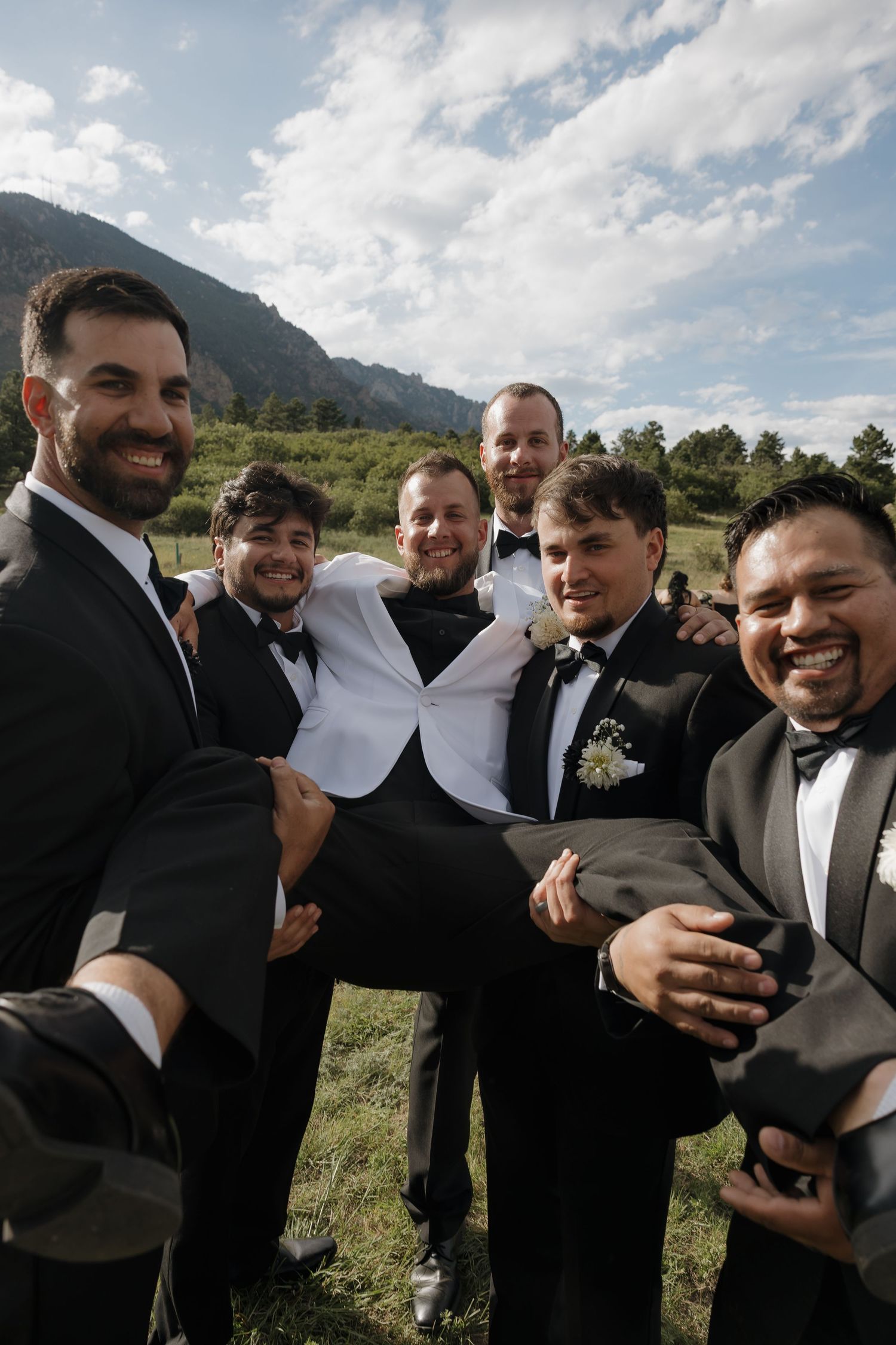 Groom in white tuxedo lifted by smiling groomsmen in black suits with mountain scenery behind.