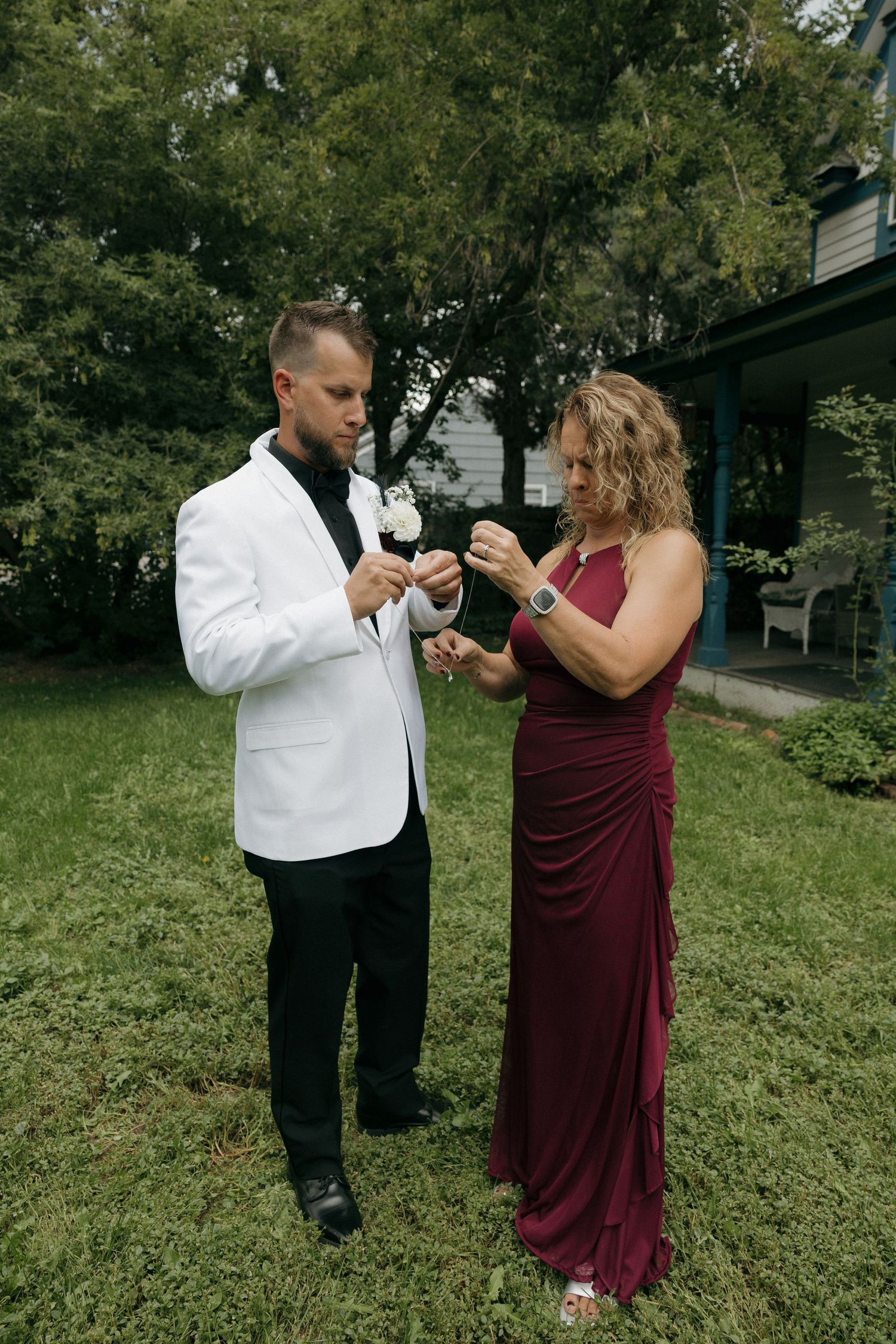 Groom in white blazer receiving boutonniere from woman in burgundy gown outdoors.
