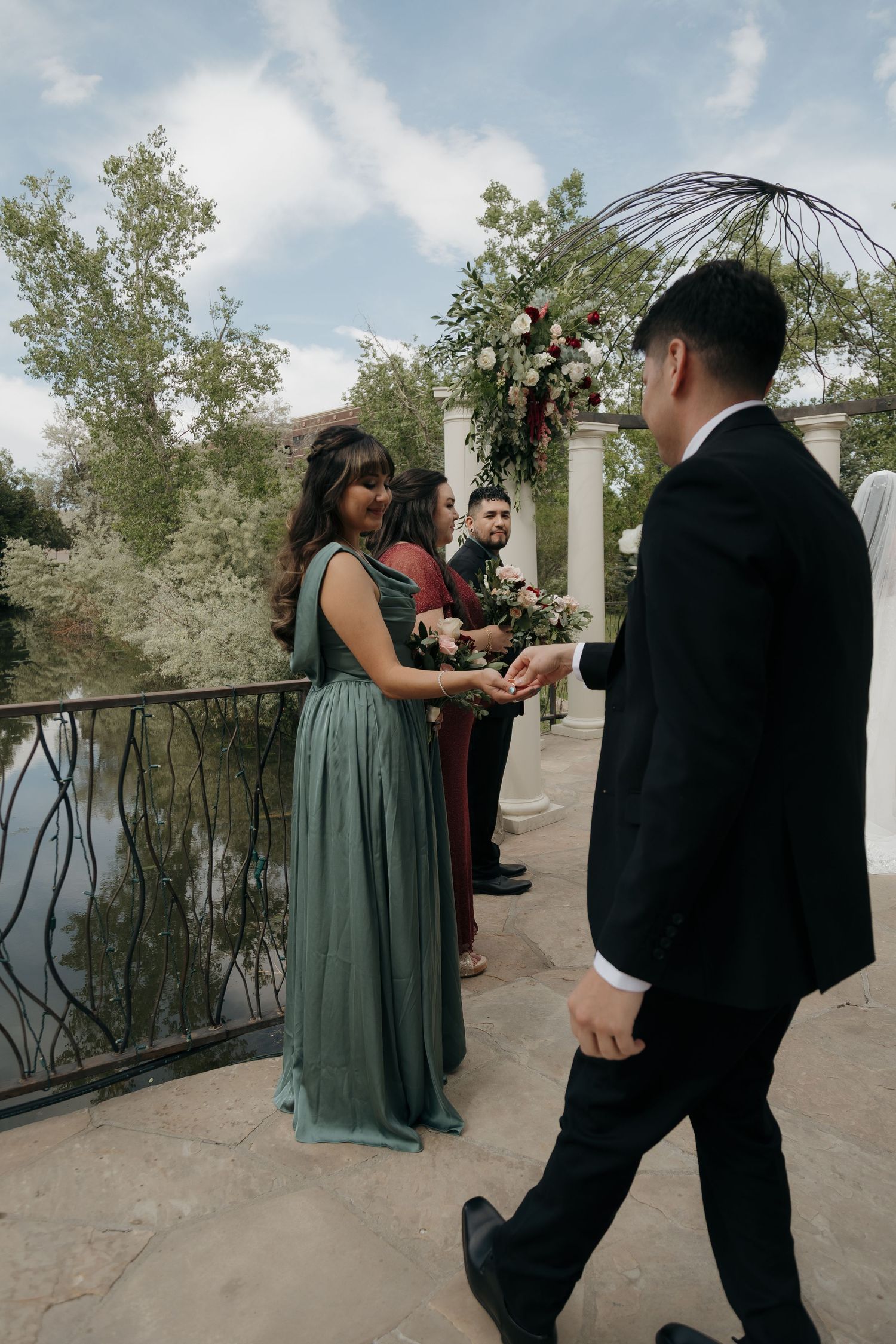 Groomsman escorts bridesmaid in green gown at outdoor wedding ceremony by a pond.