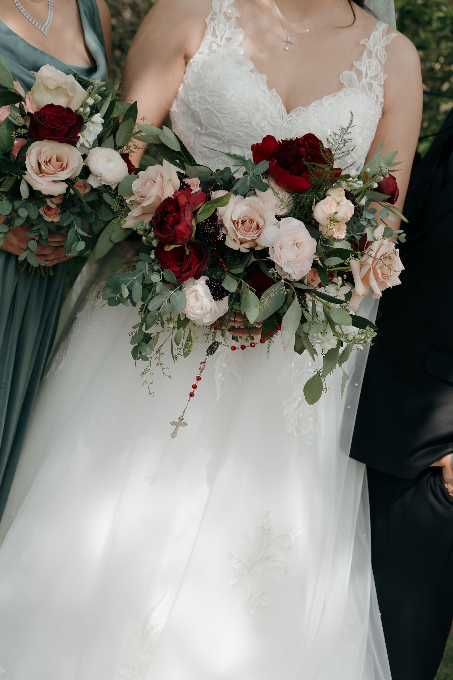 A bride in a lace wedding gown holds a bouquet of red, pink, and white roses with a rosary.