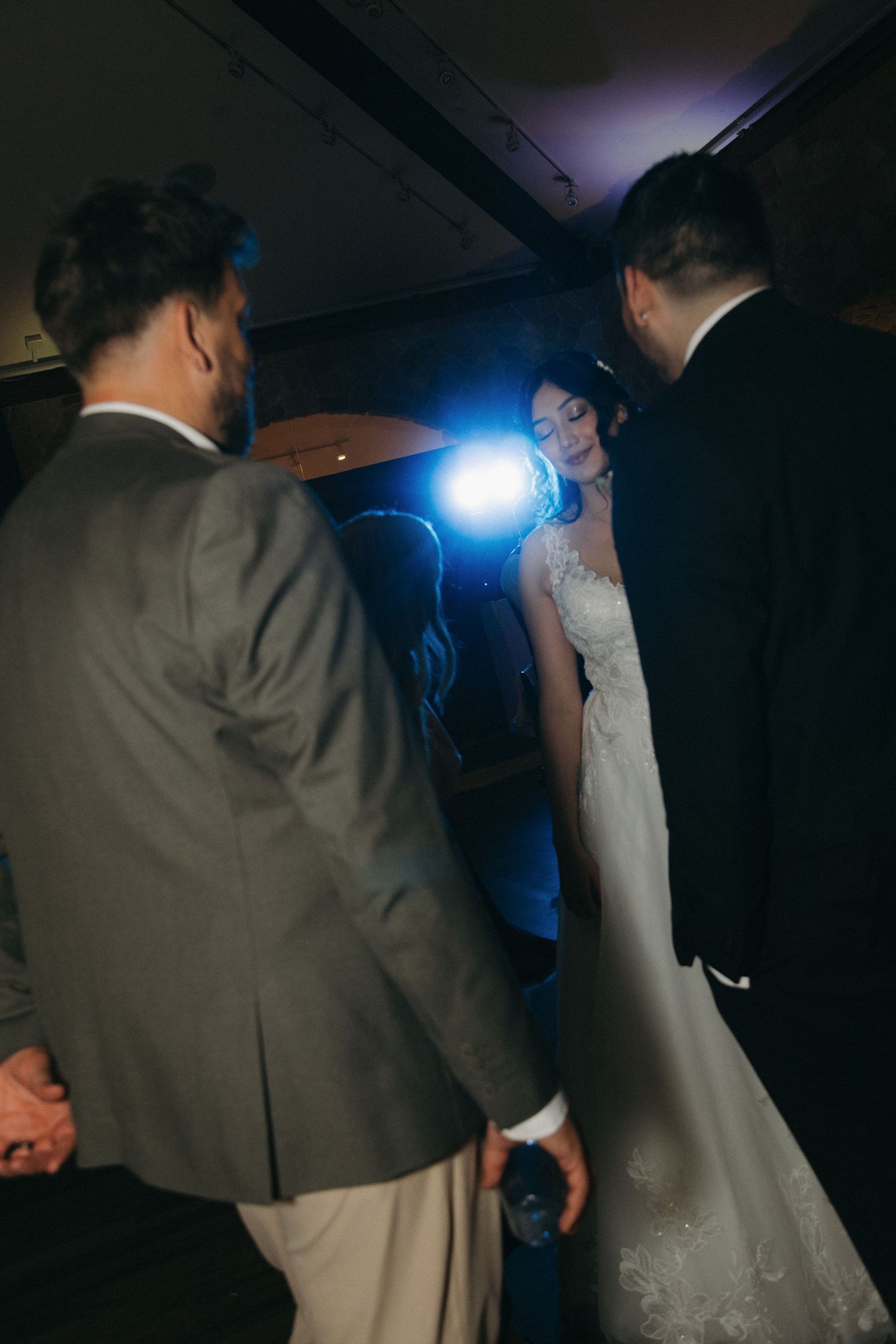 Bride in lace gown dancing with guests under a bright spotlight at a dark wedding reception.