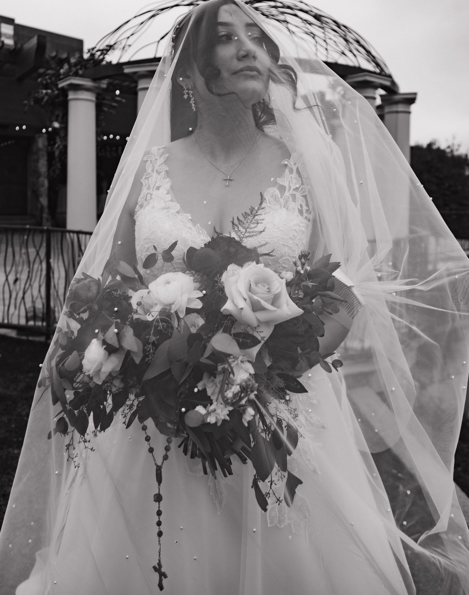 Bride in lace wedding gown holds floral bouquet with roses under flowing veil in black and white portrait.