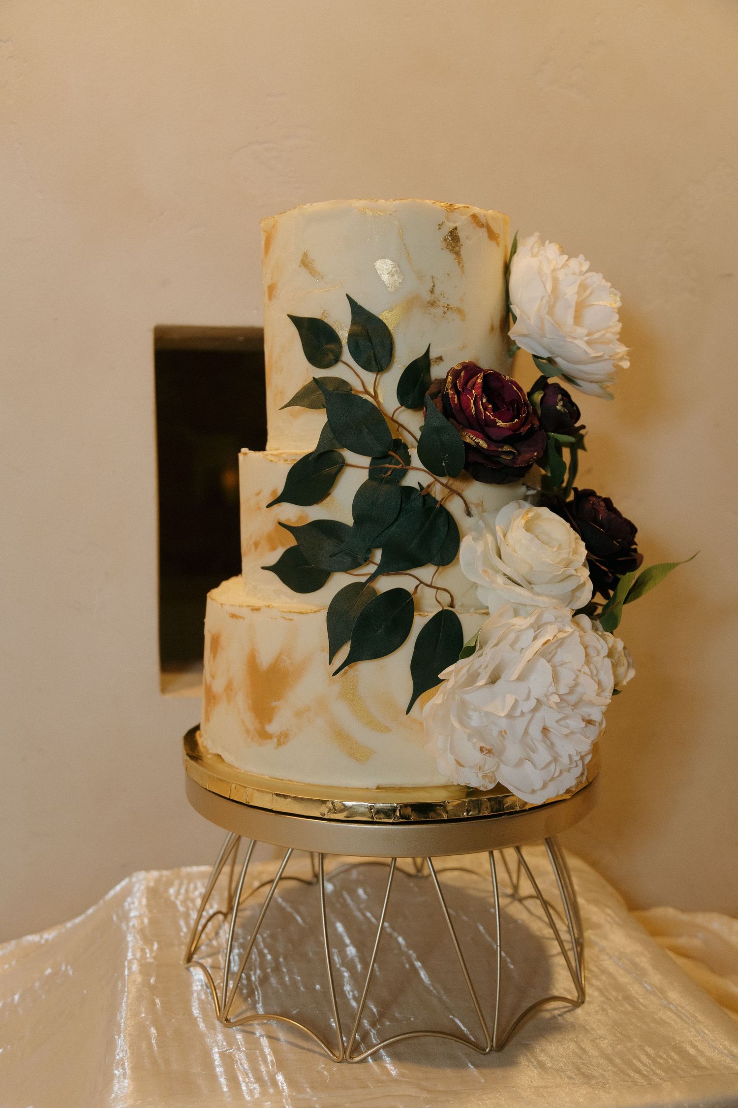Elegant two-tier white wedding cake with gold accents, dark roses, and white peonies on a geometric gold stand.