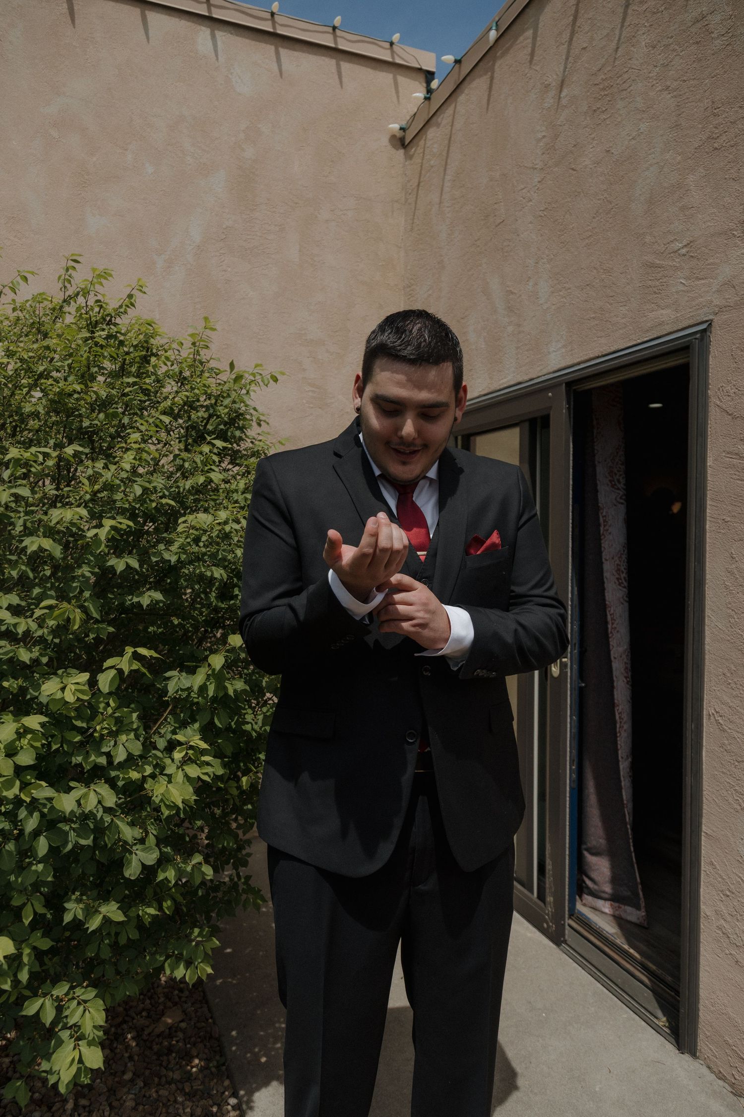 Groom in dark suit with red tie adjusting cufflinks outdoors near a beige stucco building.
