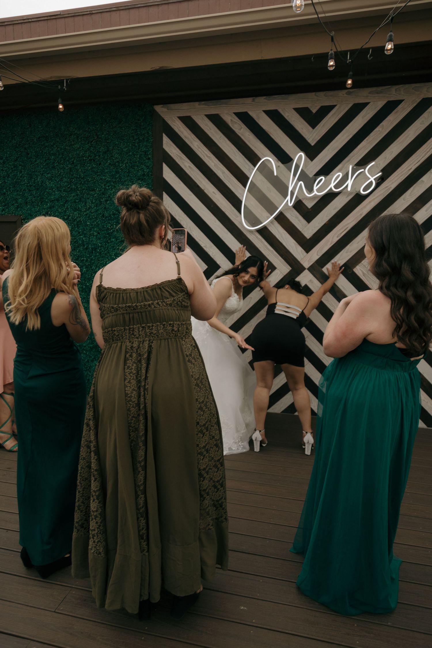 Wedding guests photographing bride near geometric Cheers sign at outdoor rooftop reception.
