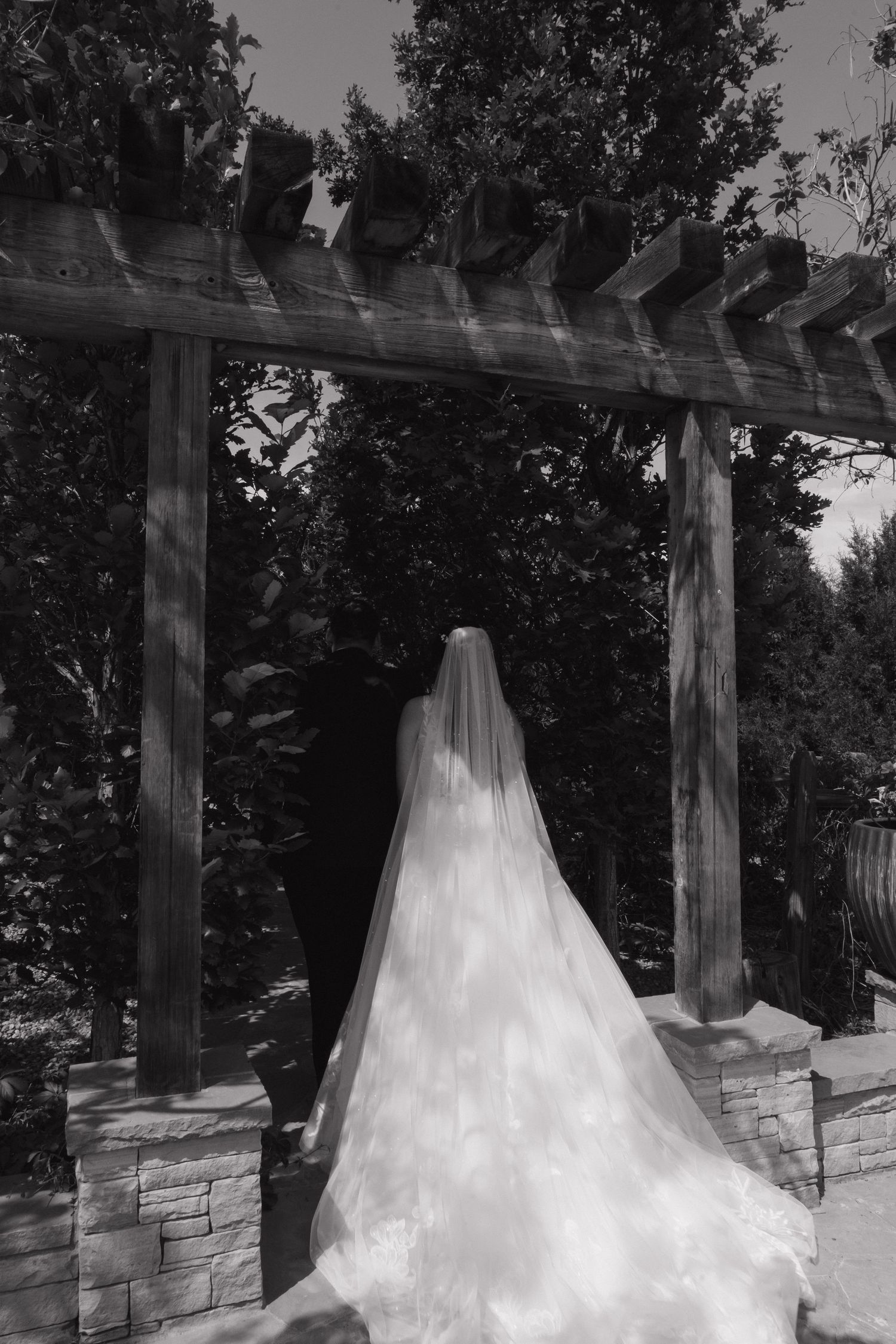 Black and white photo of bride's veil trailing under a rustic wooden pergola.