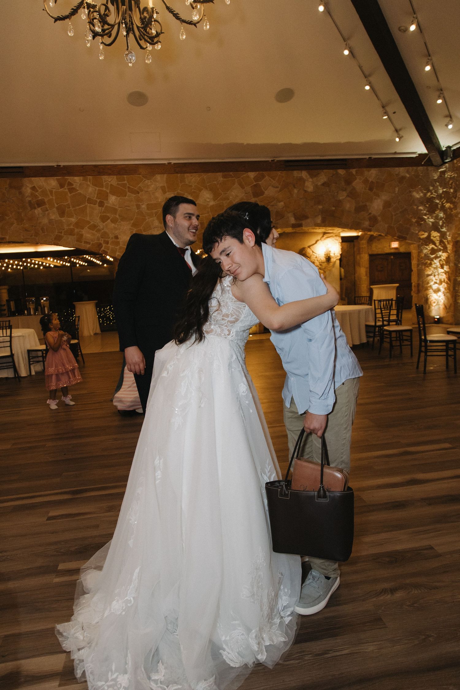 Bride in white gown receives a hug from young guest on the wedding reception dance floor.