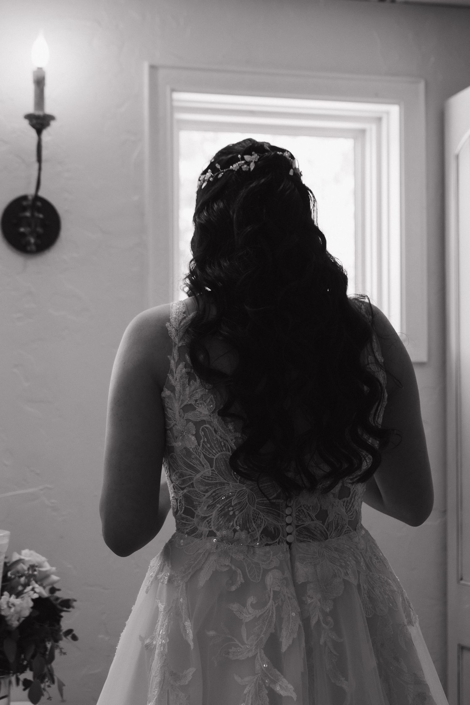 Black and white photo of bride in lace wedding gown with floral hair accessory standing by window.