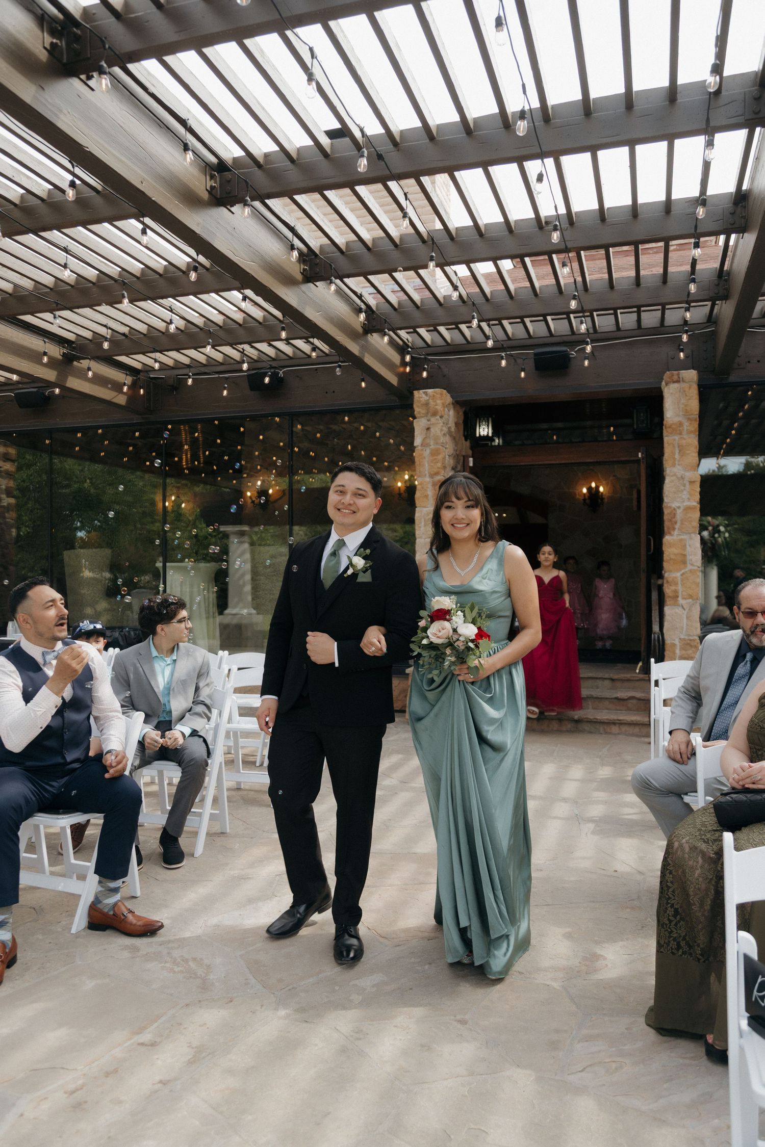 Bridesmaid in sage green gown walks down wedding aisle with groomsman under string lights at elegant indoor venue.