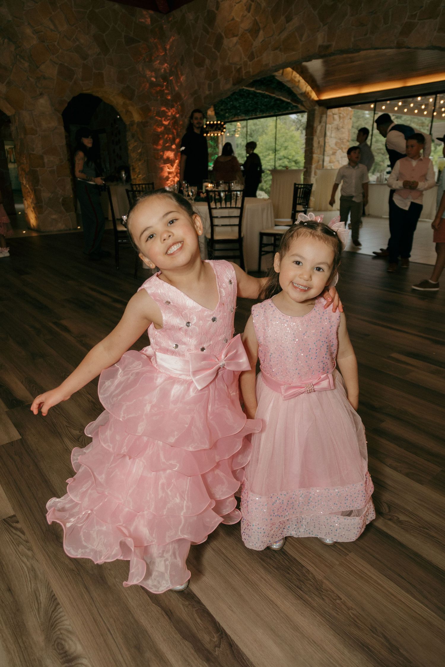 Two young flower girls in matching pink ruffled dresses smiling and dancing at wedding reception.