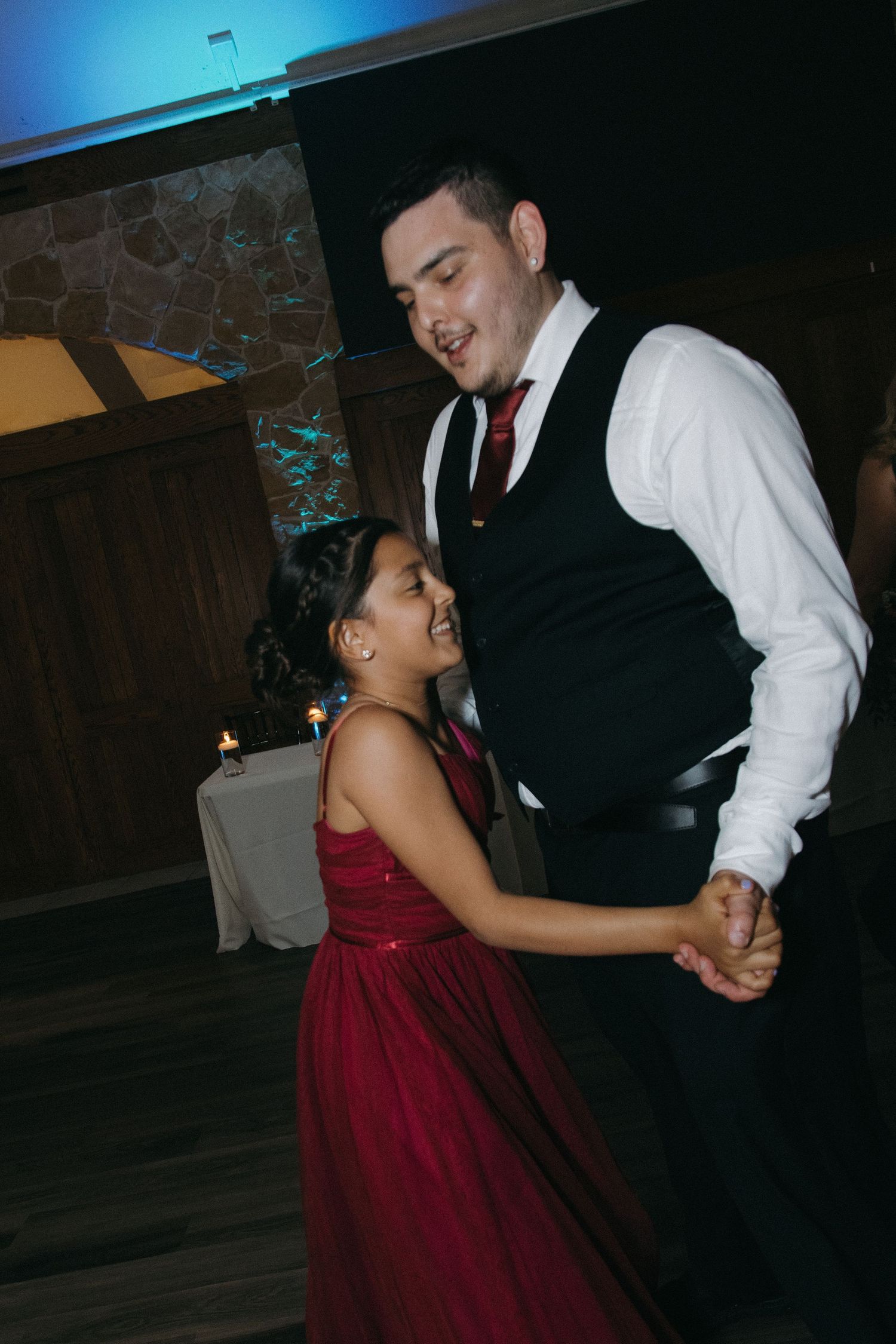 Joyful man in vest and red tie dances with a young girl in a red dress at a wedding reception.