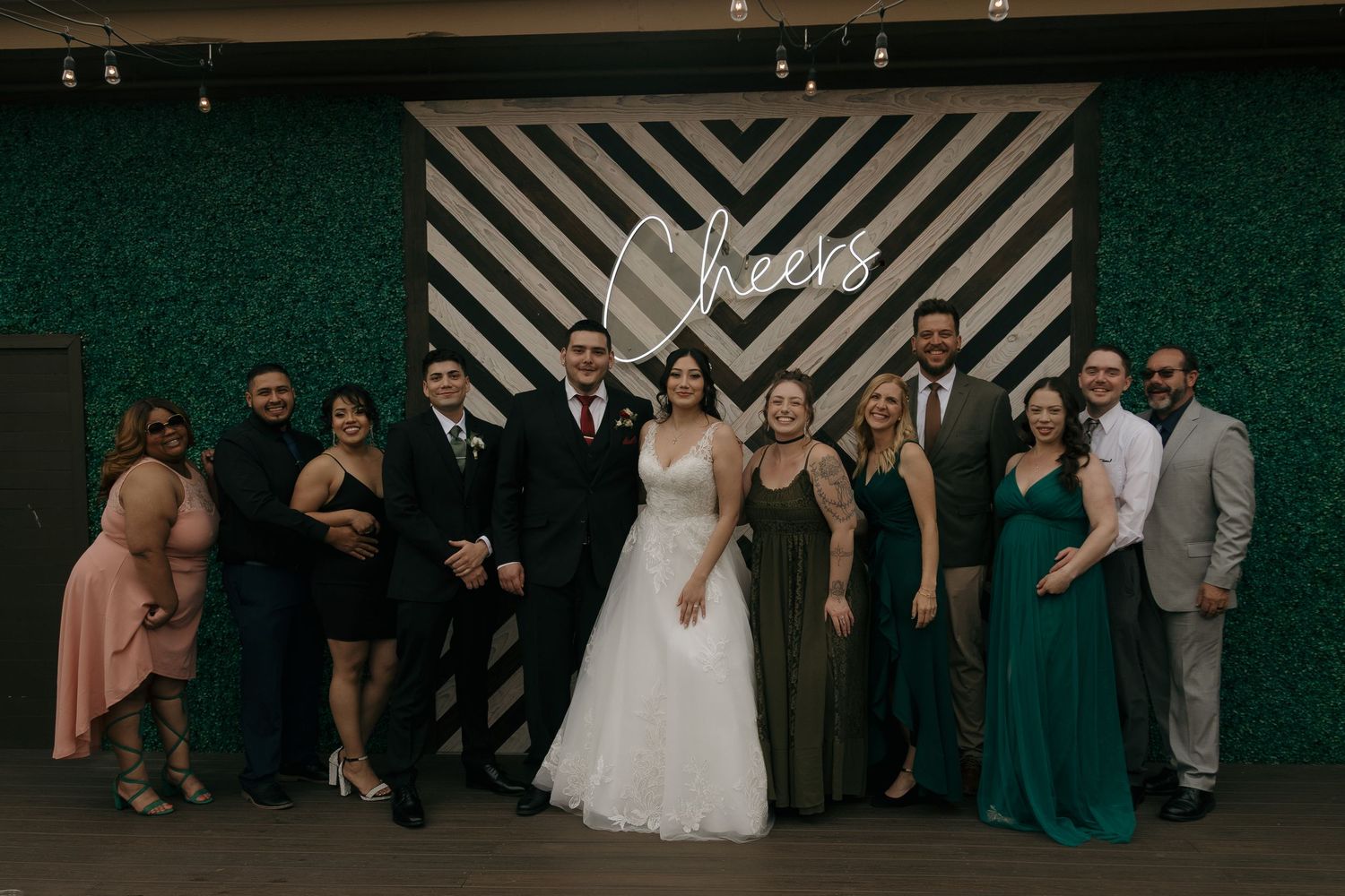 Wedding party poses together in front of a Cheers neon sign backdrop at the reception.