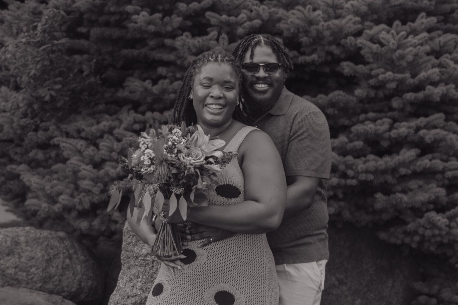 Black and white photo of a happy couple embracing outdoors with a bouquet against an evergreen backdrop.