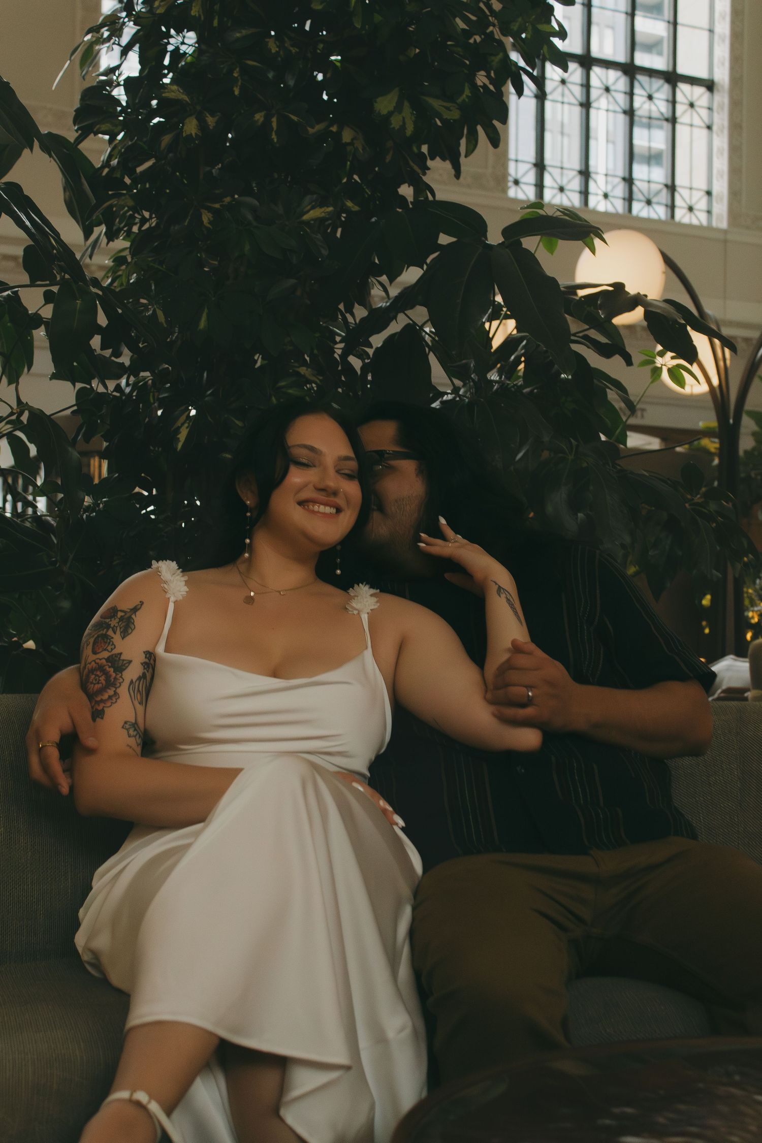 A couple shares a joyful moment together on a couch surrounded by indoor plants.