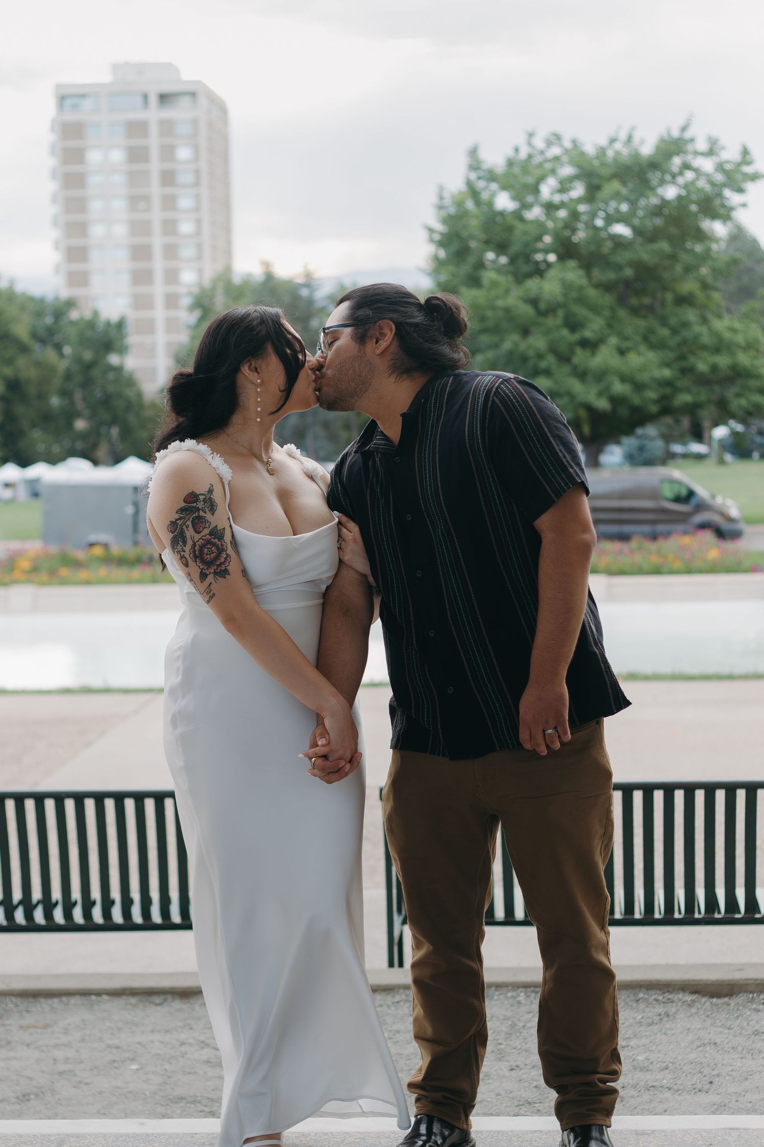 A couple shares an intimate moment on a bench with urban buildings and trees in the background.