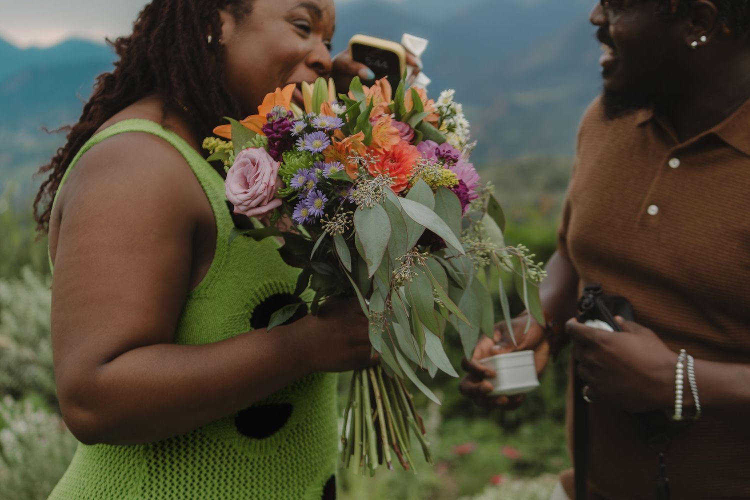 Two people share a tender moment on a mountainside while admiring a vibrant bouquet of mixed flowers.
