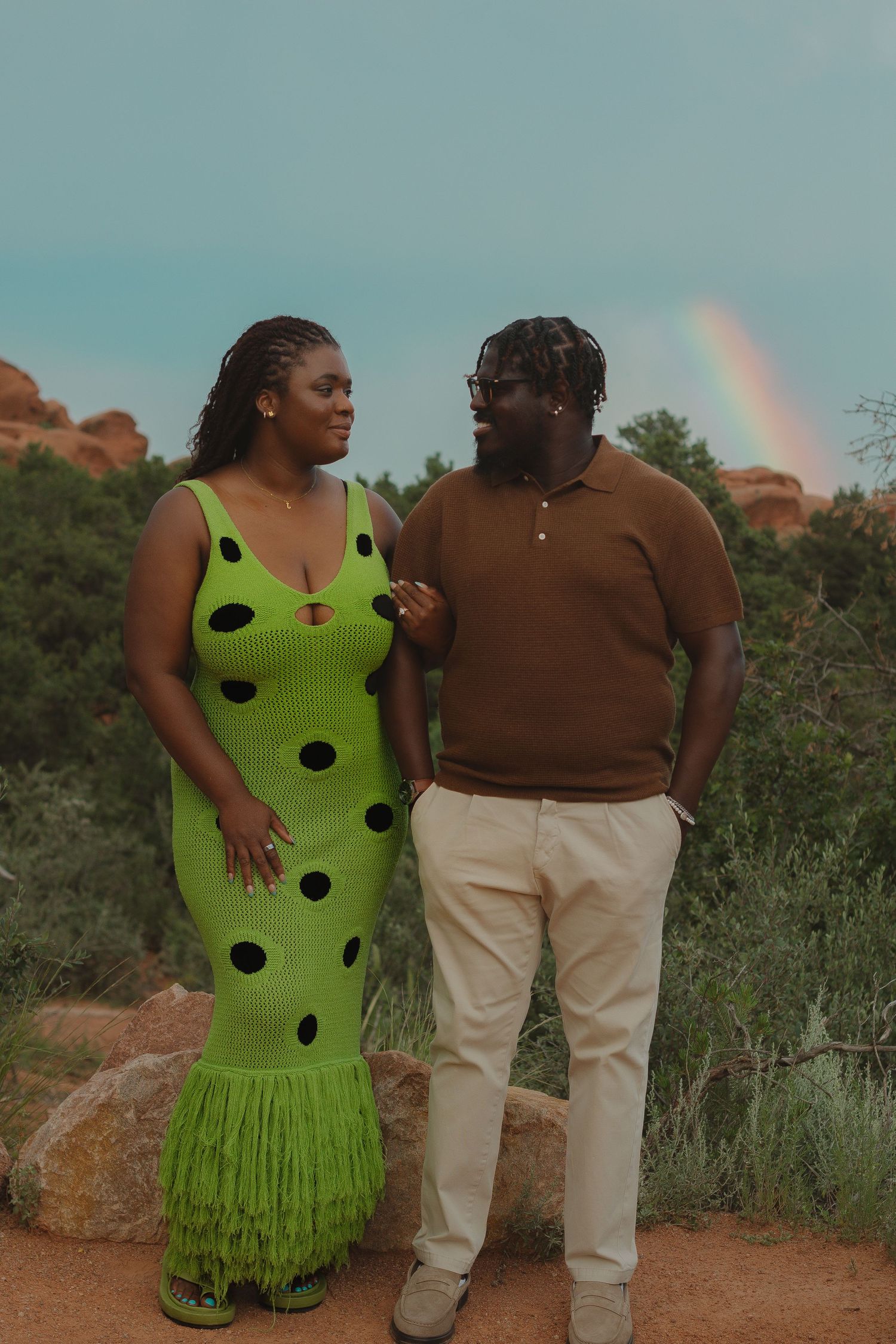 Couple poses together on a nature trail, one in a vibrant lime green polka dot dress and the other in brown.