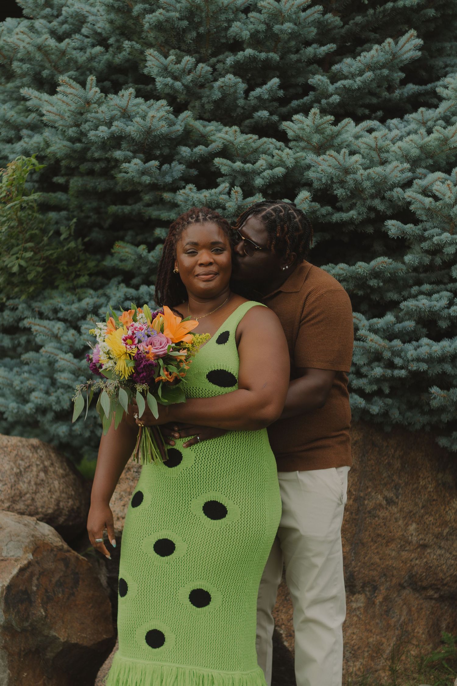 A romantic couple poses with colorful flowers against blue spruce trees, wearing lime green and brown outfits.