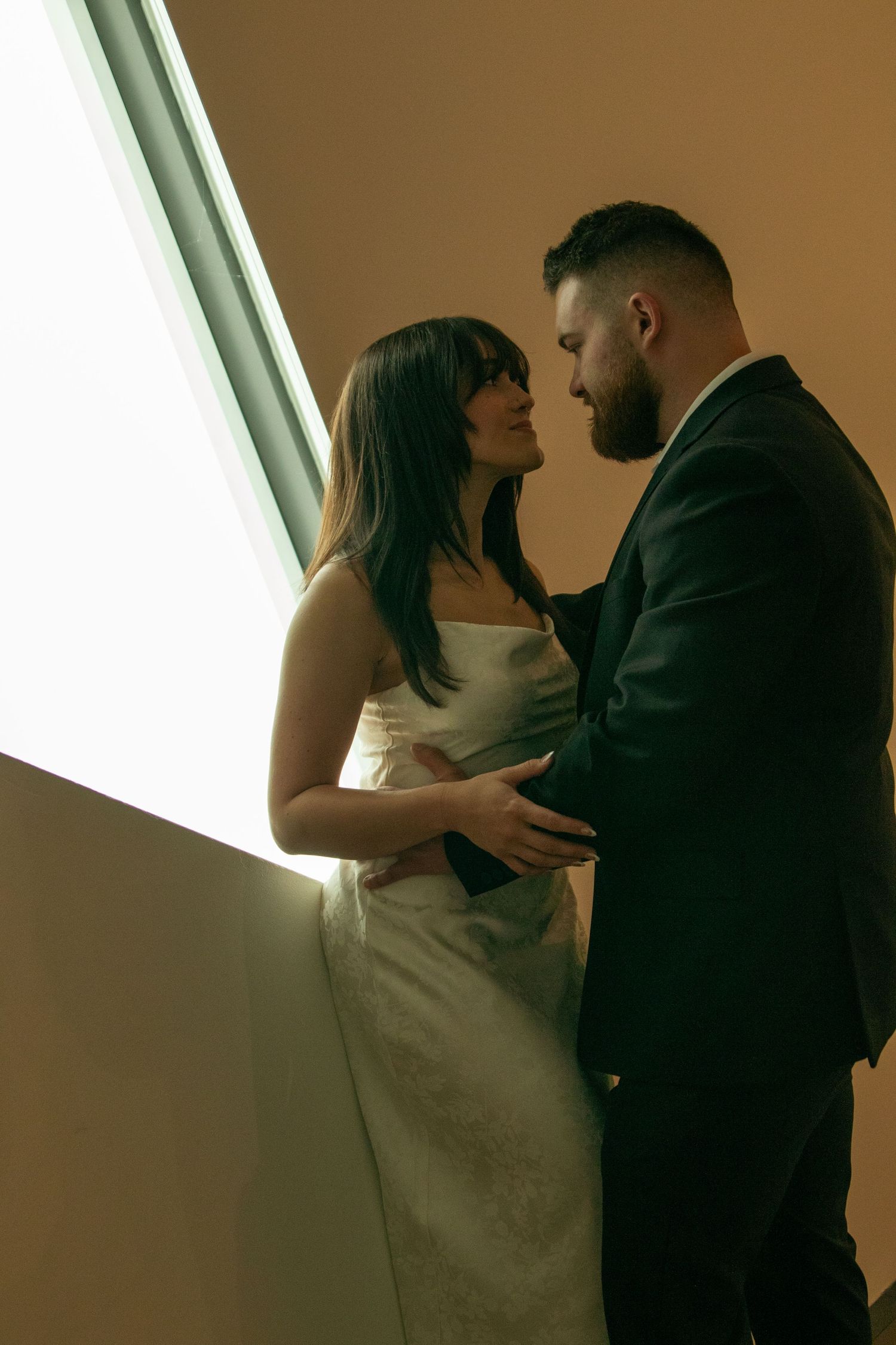 Silhouetted couple in formal attire dancing intimately near a bright window in warm lighting.