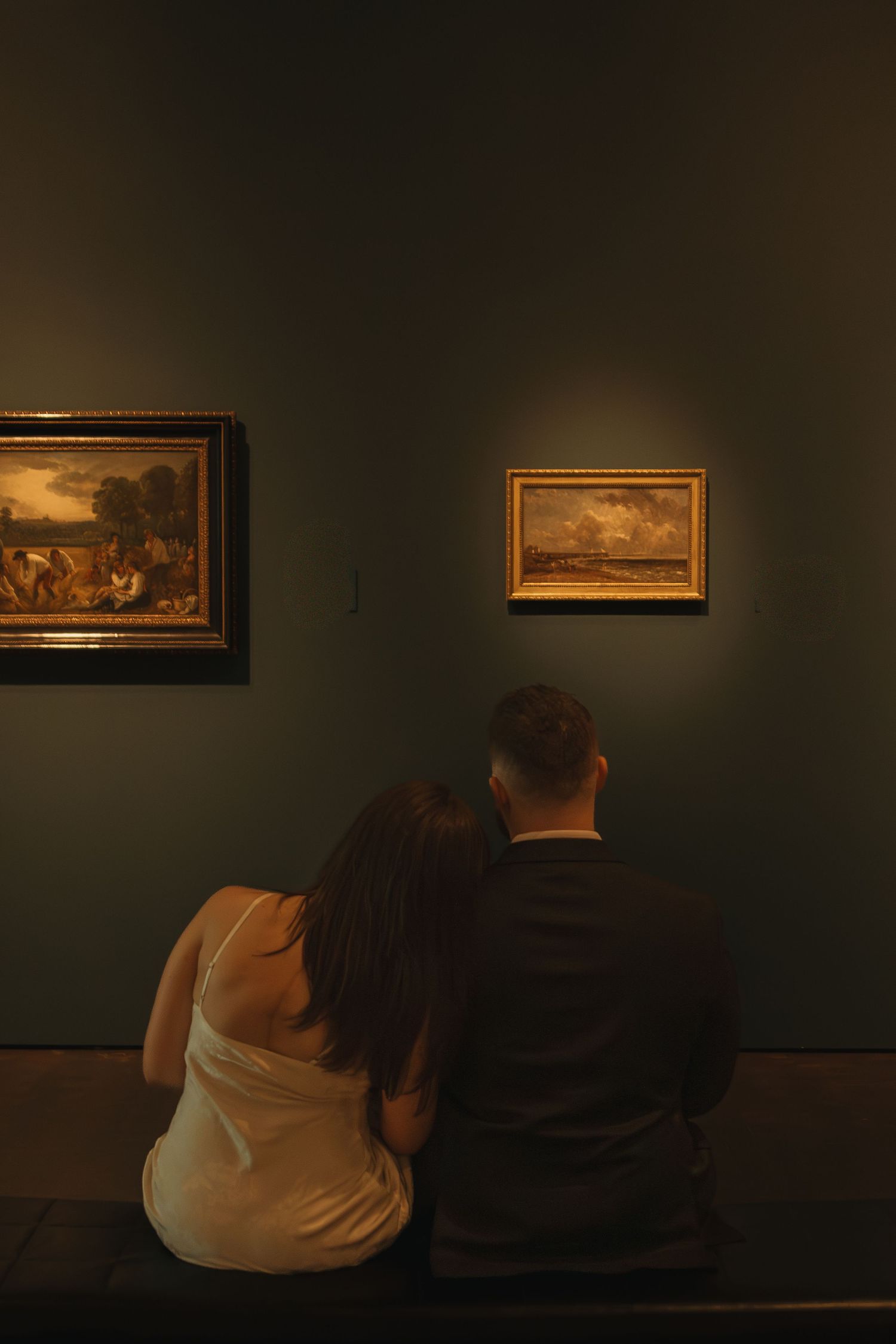 Two people sit close together viewing classical paintings in a dark museum gallery.