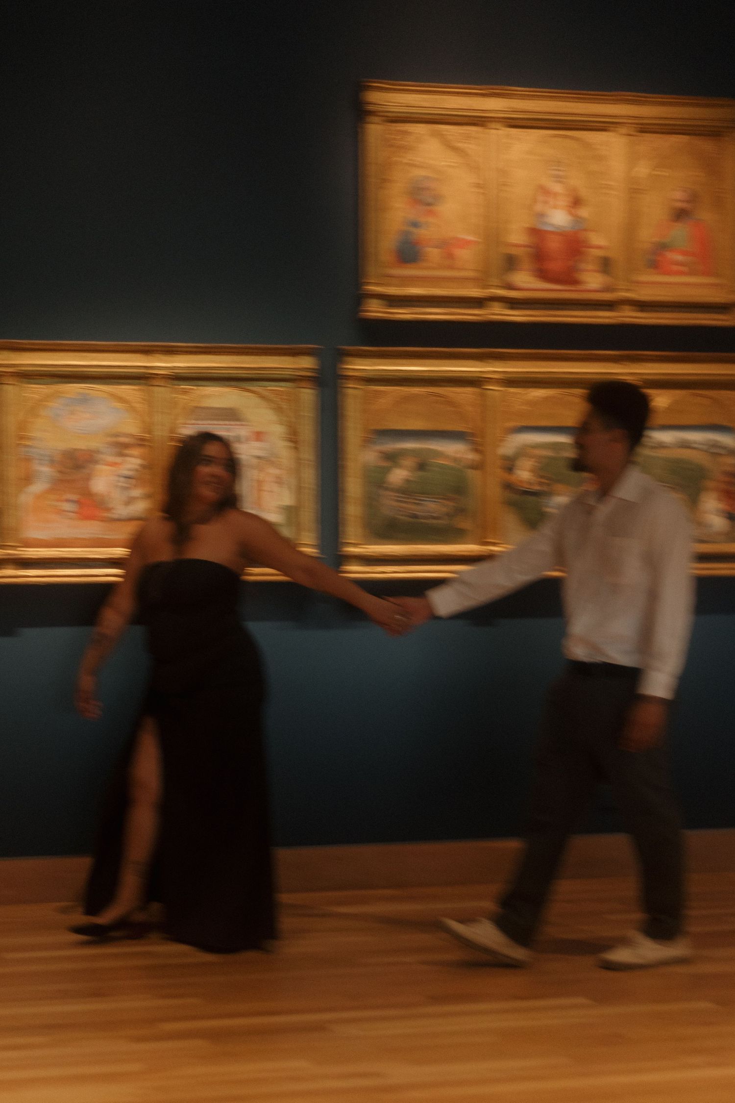 Couple holding hands in a museum gallery with ornate gold-framed Renaissance paintings on teal walls.