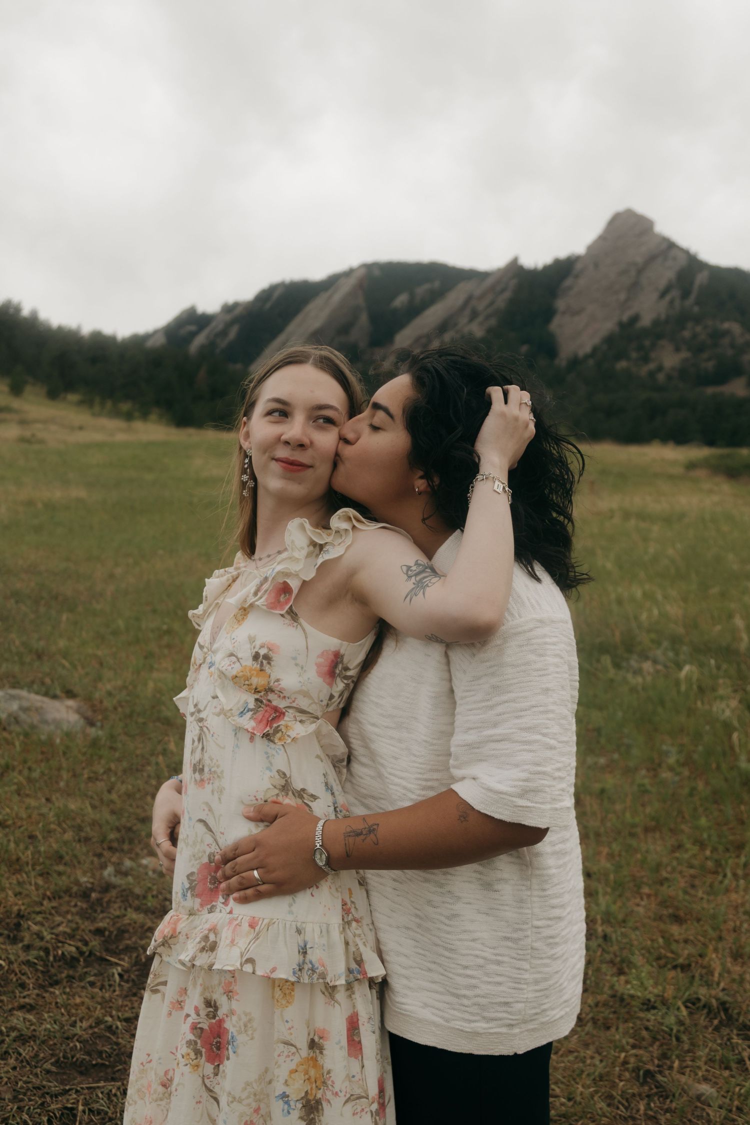 Two women embracing in a field with rocky Flatiron mountains behind them on a cloudy day.